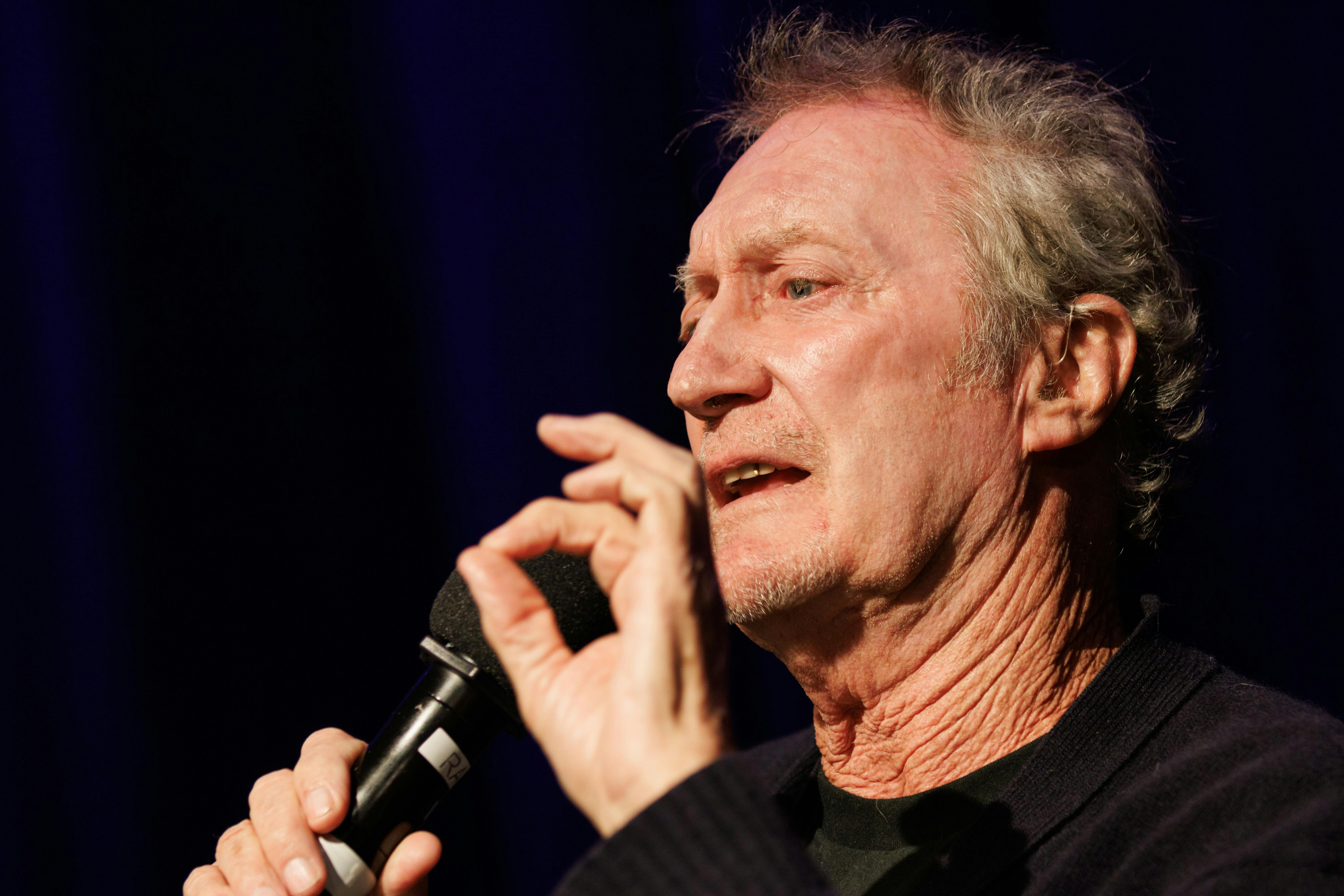 Actor Bryan Brown with greying hair holds a microphone in his right hand and is speaking.