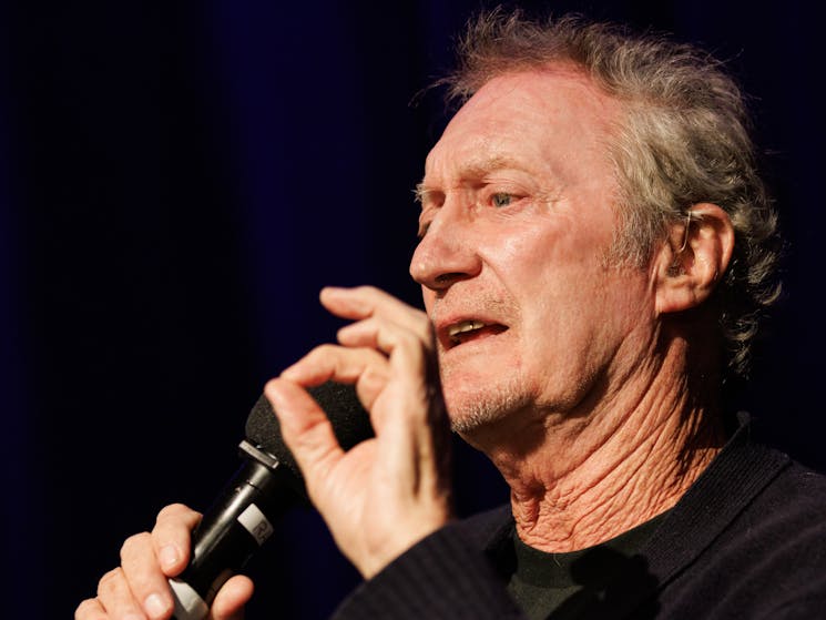 Actor Bryan Brown with greying hair holds a microphone in his right hand and is speaking.
