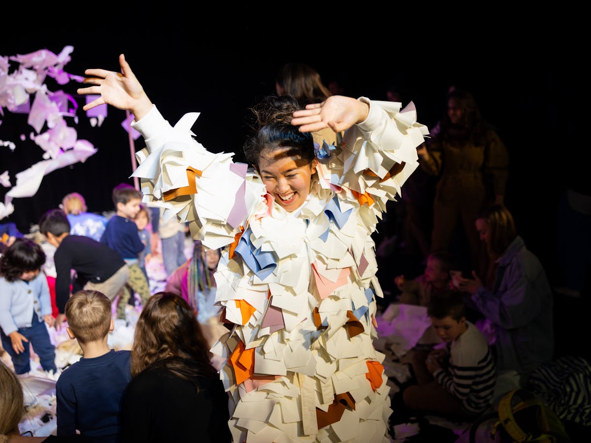 Performance documentation of Club Origami. Perfomer is smiling with hands up and paper flying around