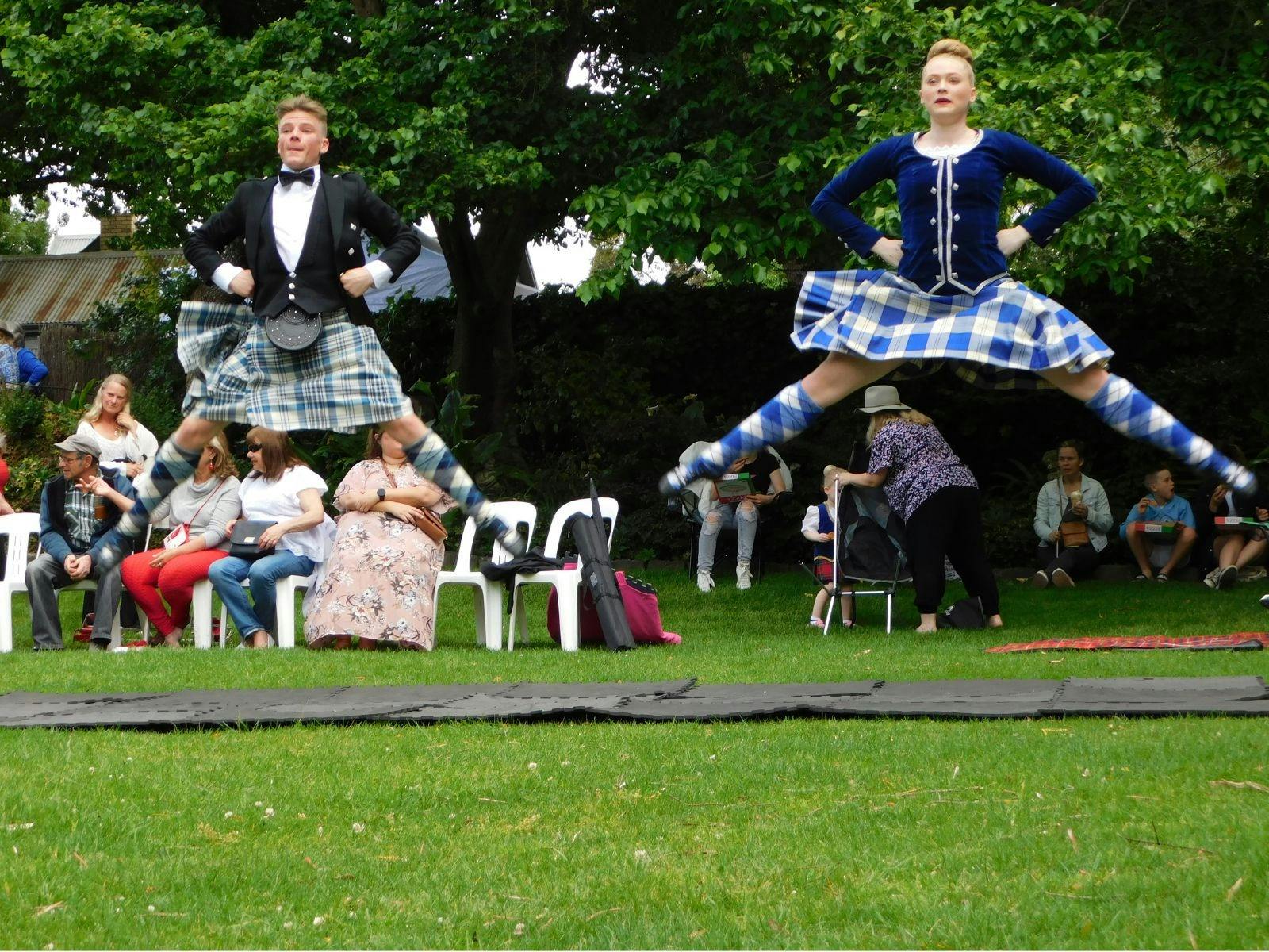 Two male dancers in mid air, wearing Scottish traditional clothing, as people watch them from behind