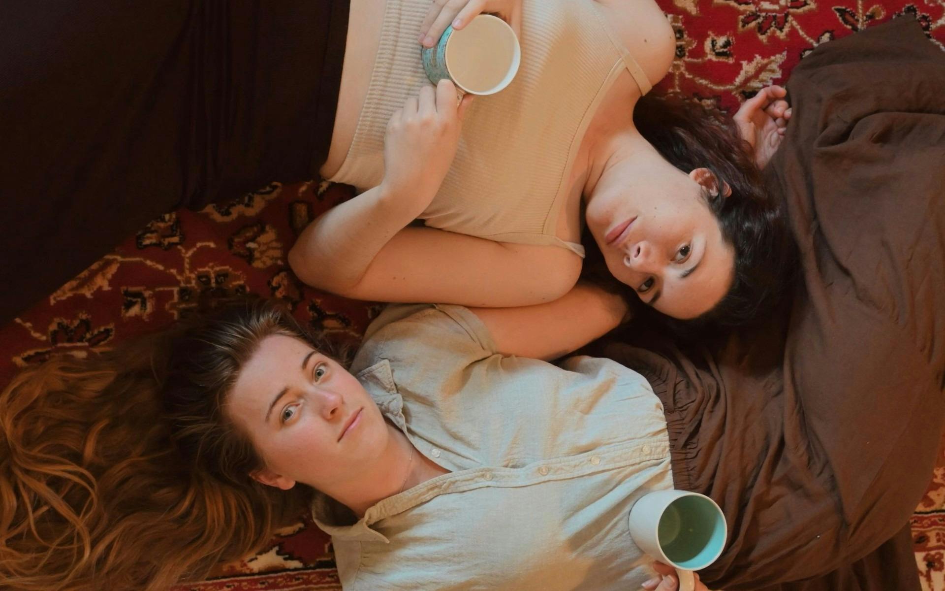 Two female actors lay parallel on a bed, each looking up at the camera with a neutral gaze.