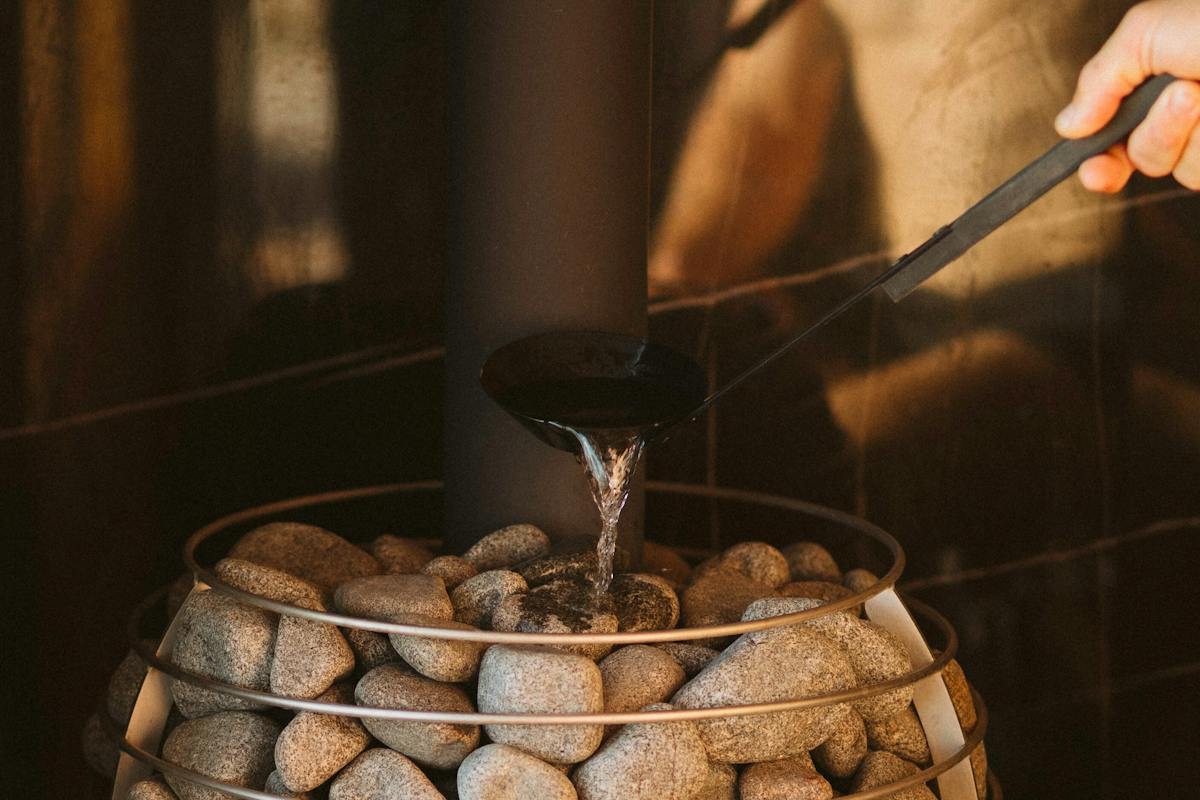 A male figure using the sauna ladle to pour water on the hot sauna rocks creating sauna steam