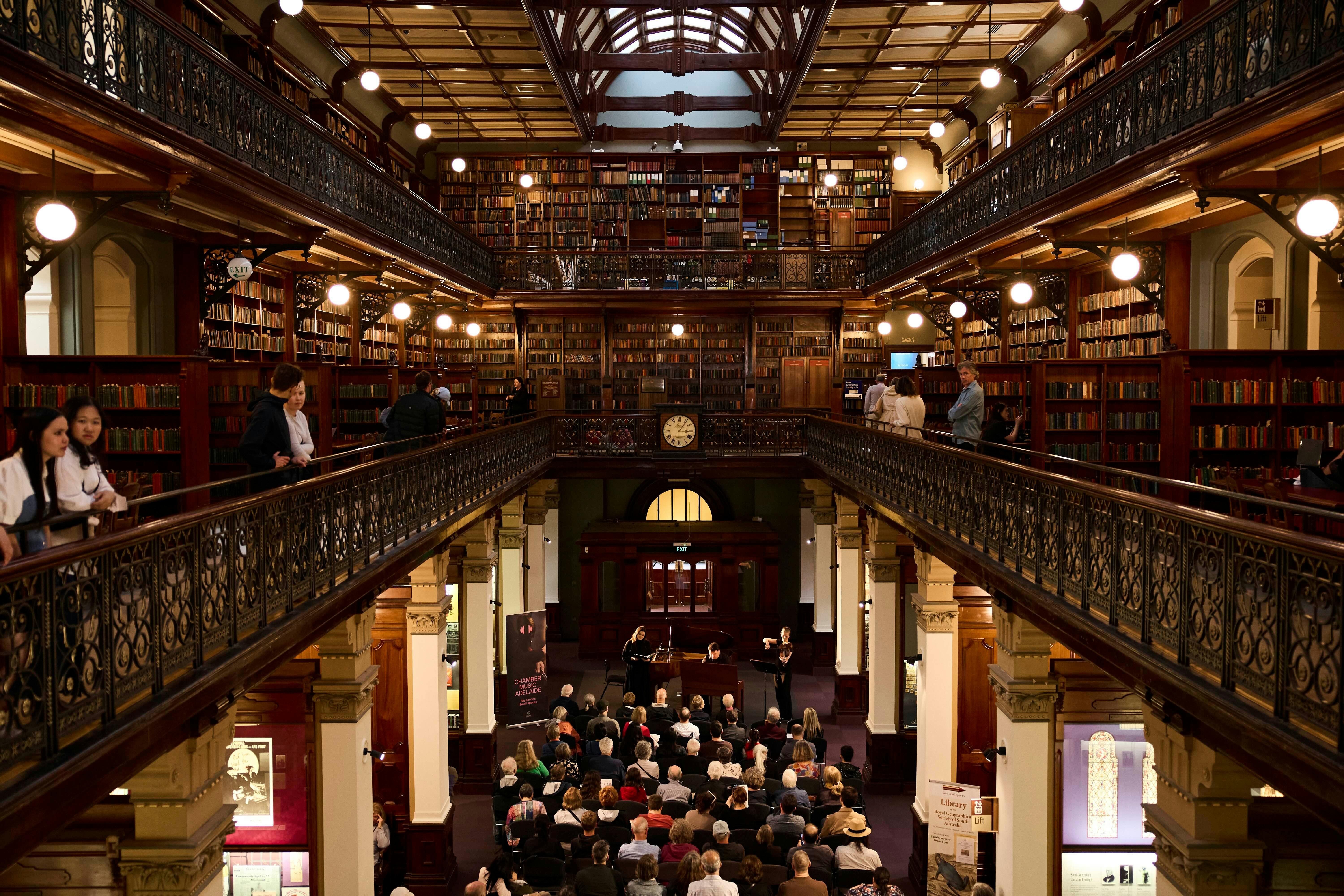 A crowd watches a music concert in an ornate library while other watch from mezzanine levels above.