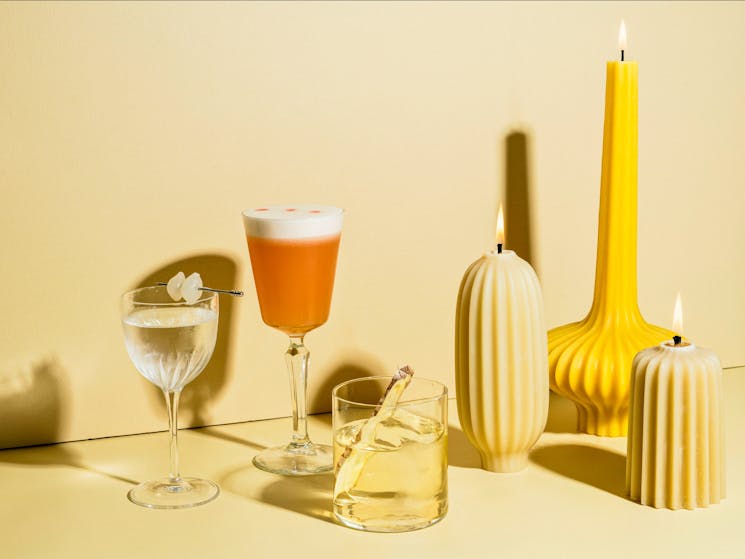 3 cocktails and 3 burning yellow candles all of various sizes sit in front of a yellow background.