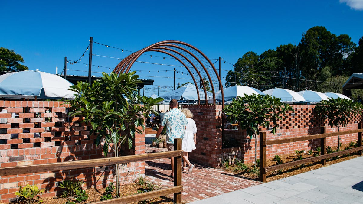 A couple walks through a brick archway entrance into a sunlit outdoor patio area with umbrellas