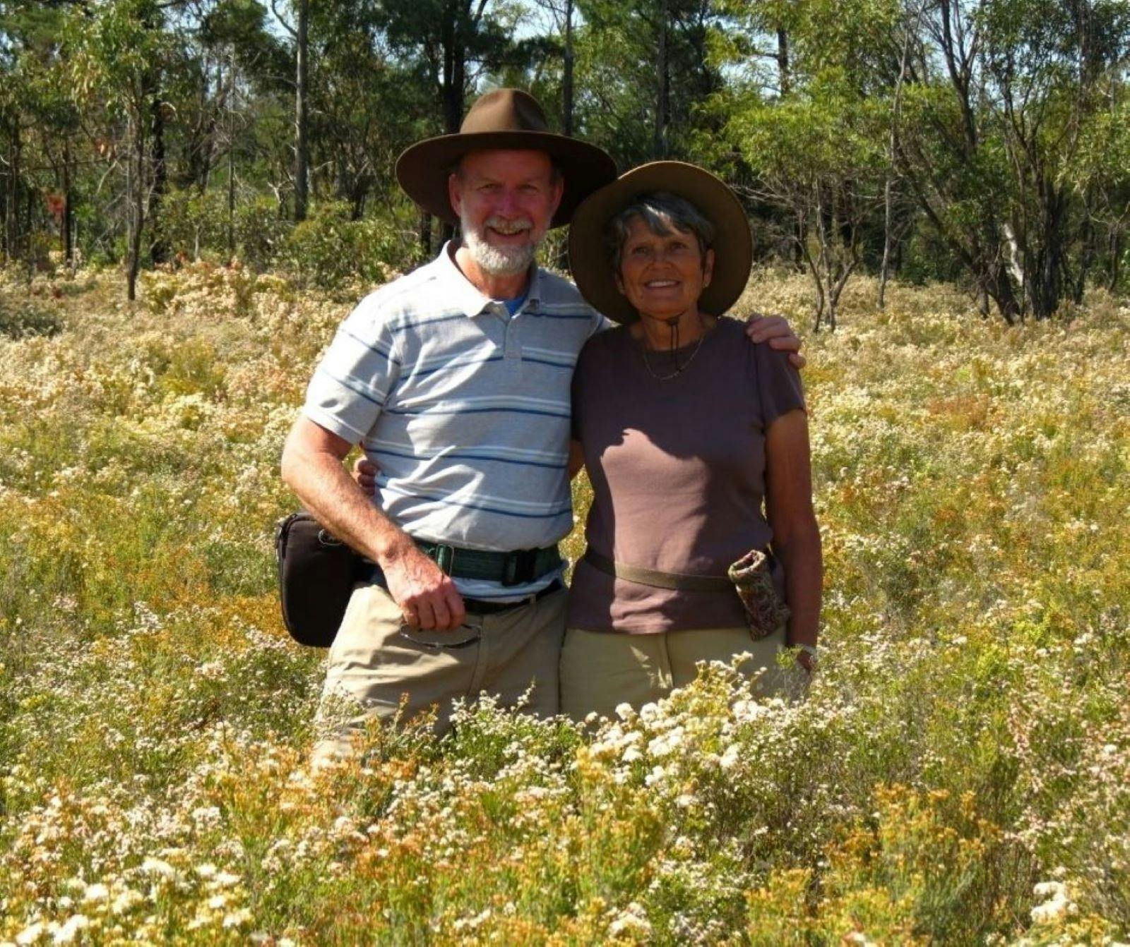 An elderly couple standing in tall grass surrounded by flowers