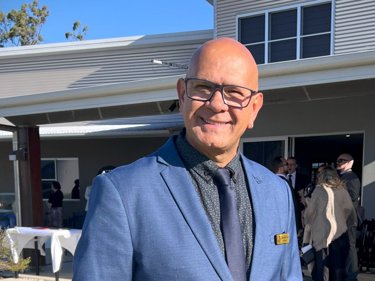 A man in a blue suit and tie stands outdoors, smiling warmly in front of a modern building during a