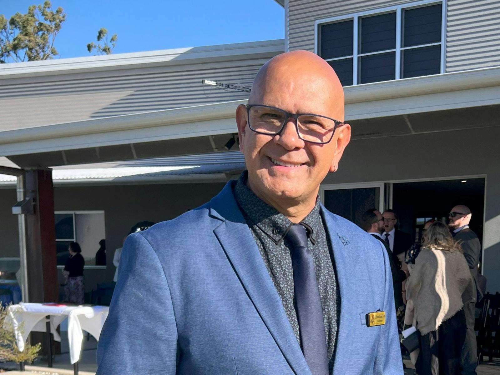 A man in a blue suit and tie stands outdoors, smiling warmly in front of a modern building during a A man in a blue suit and tie stands outdoors, smiling warmly in front of a modern building during a