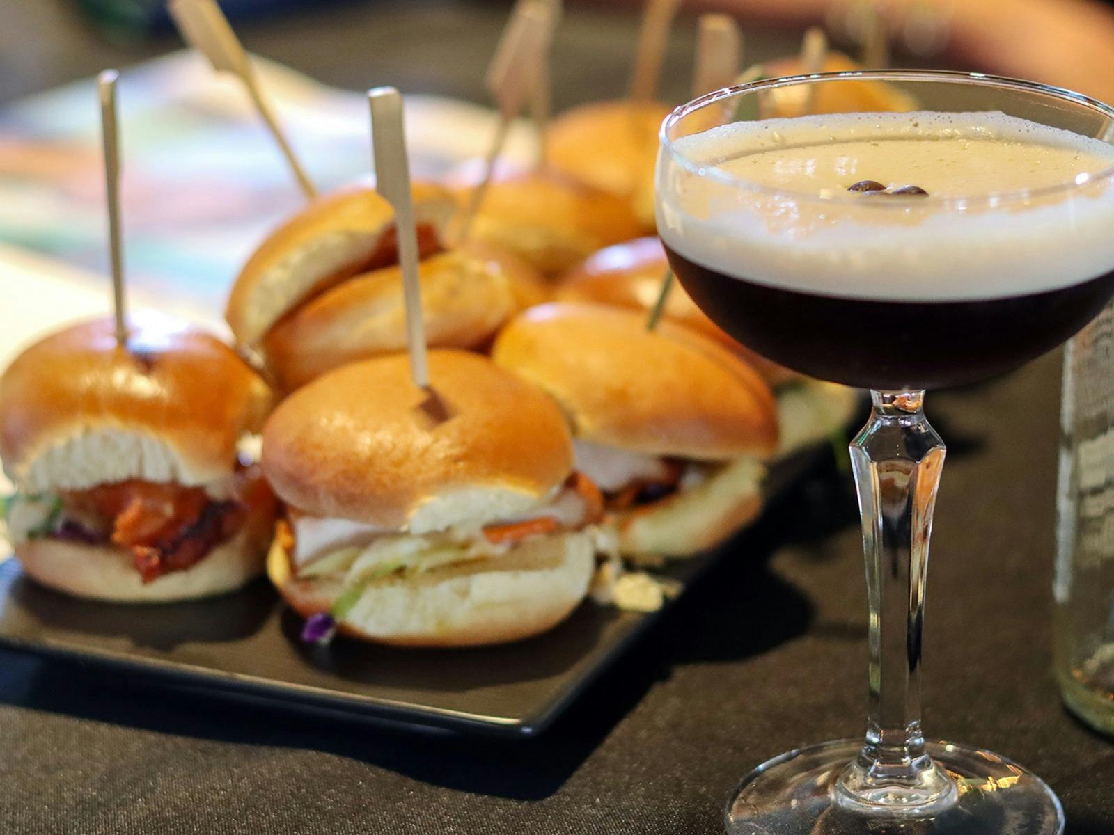 An espresso martini in the foreground with sliders on a tray in the background