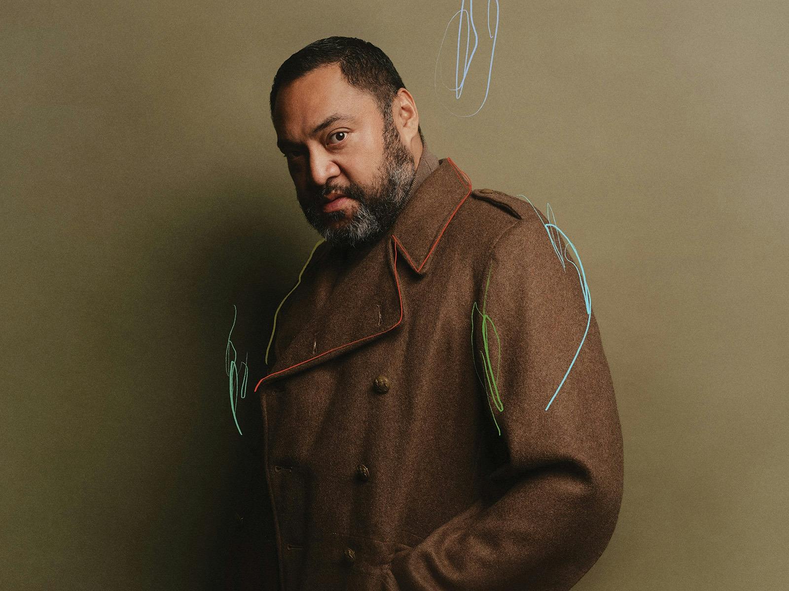 A heavy set man with a beard and dark brooding look wears a brown coat and looks directly at us