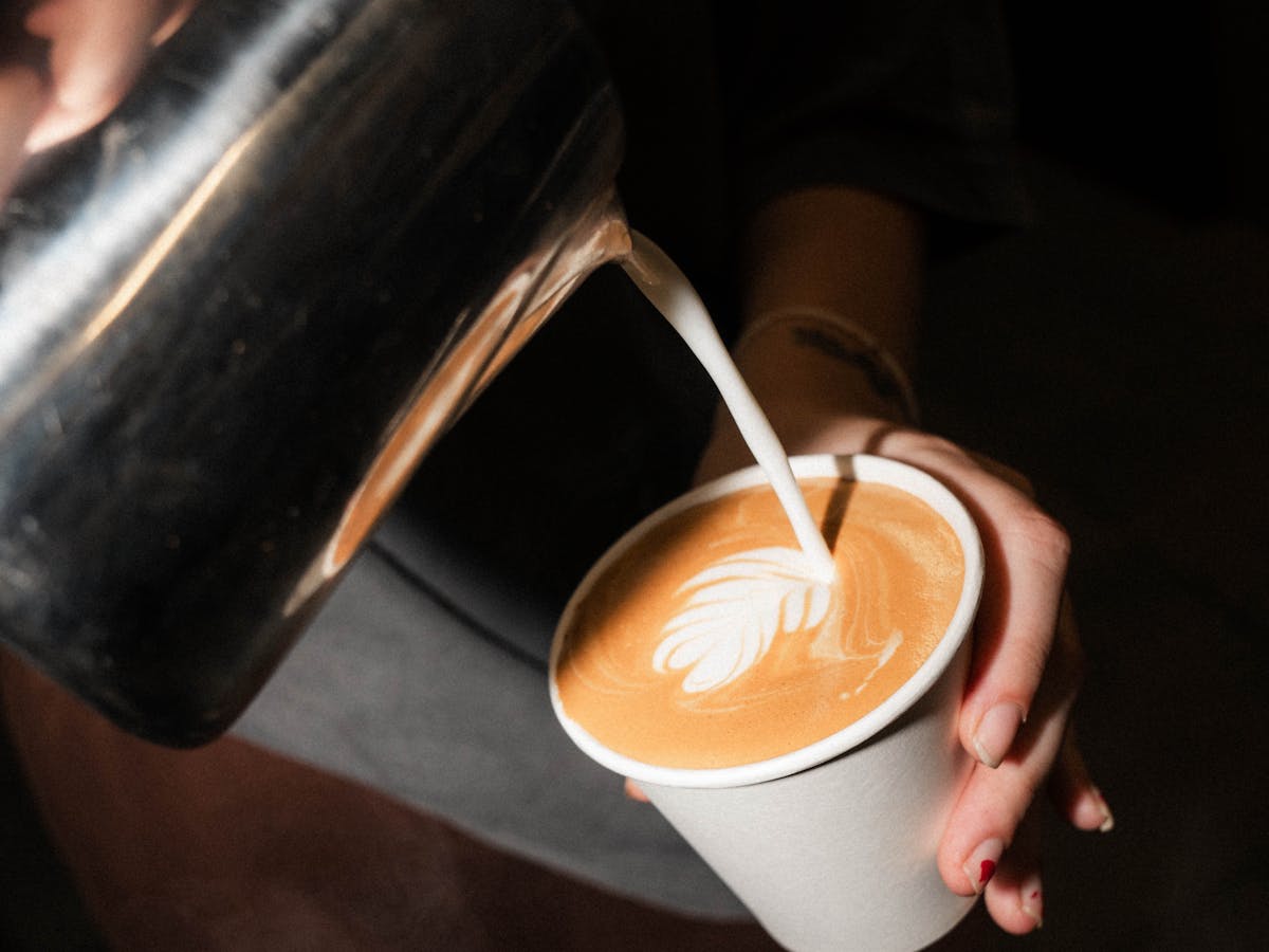 A white take away cup with milk being poured into it almost full with latte art