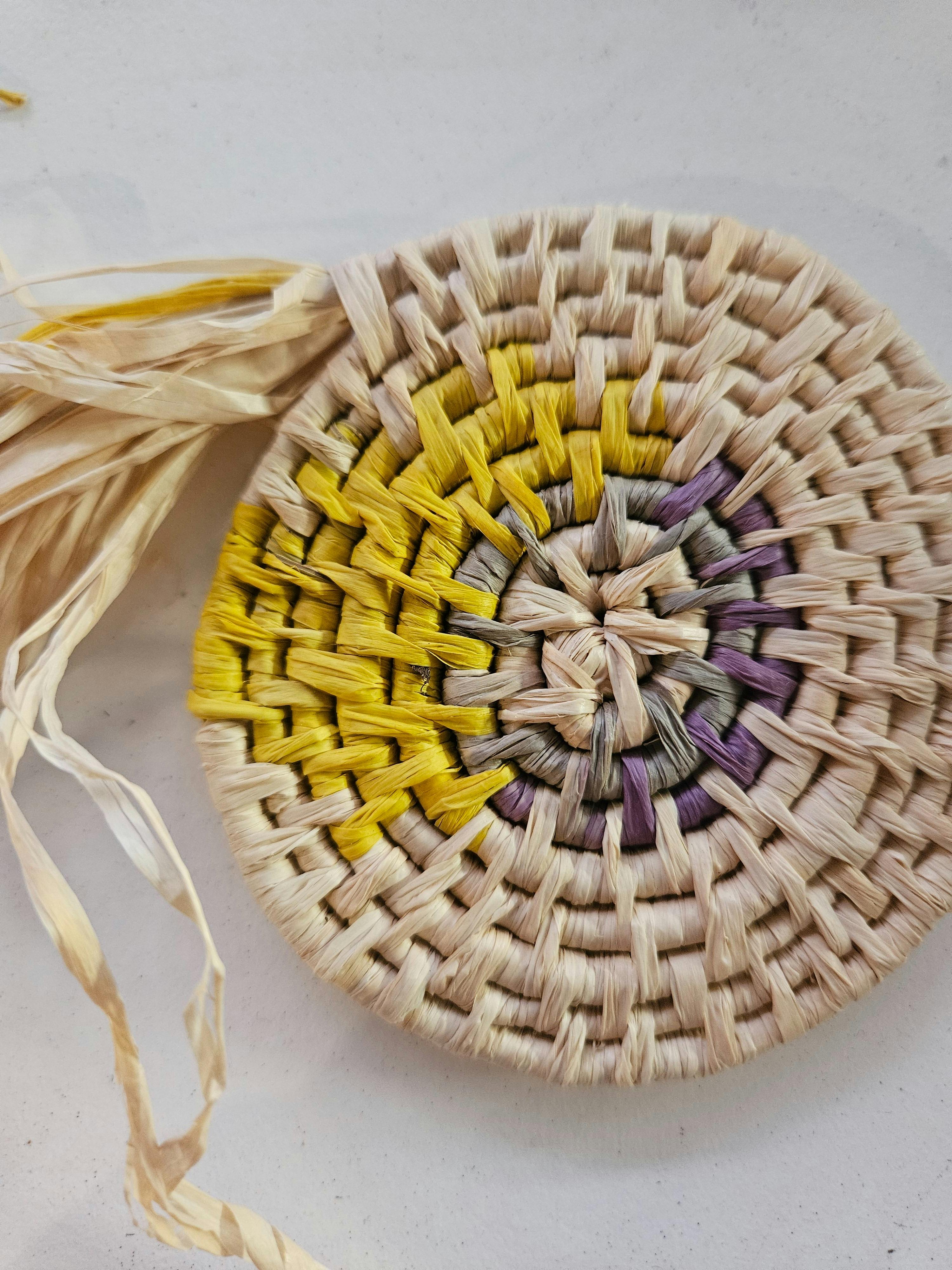 Close-up of a circular coiled raffia basketry piece featuring natural beige fibres with yellow and p