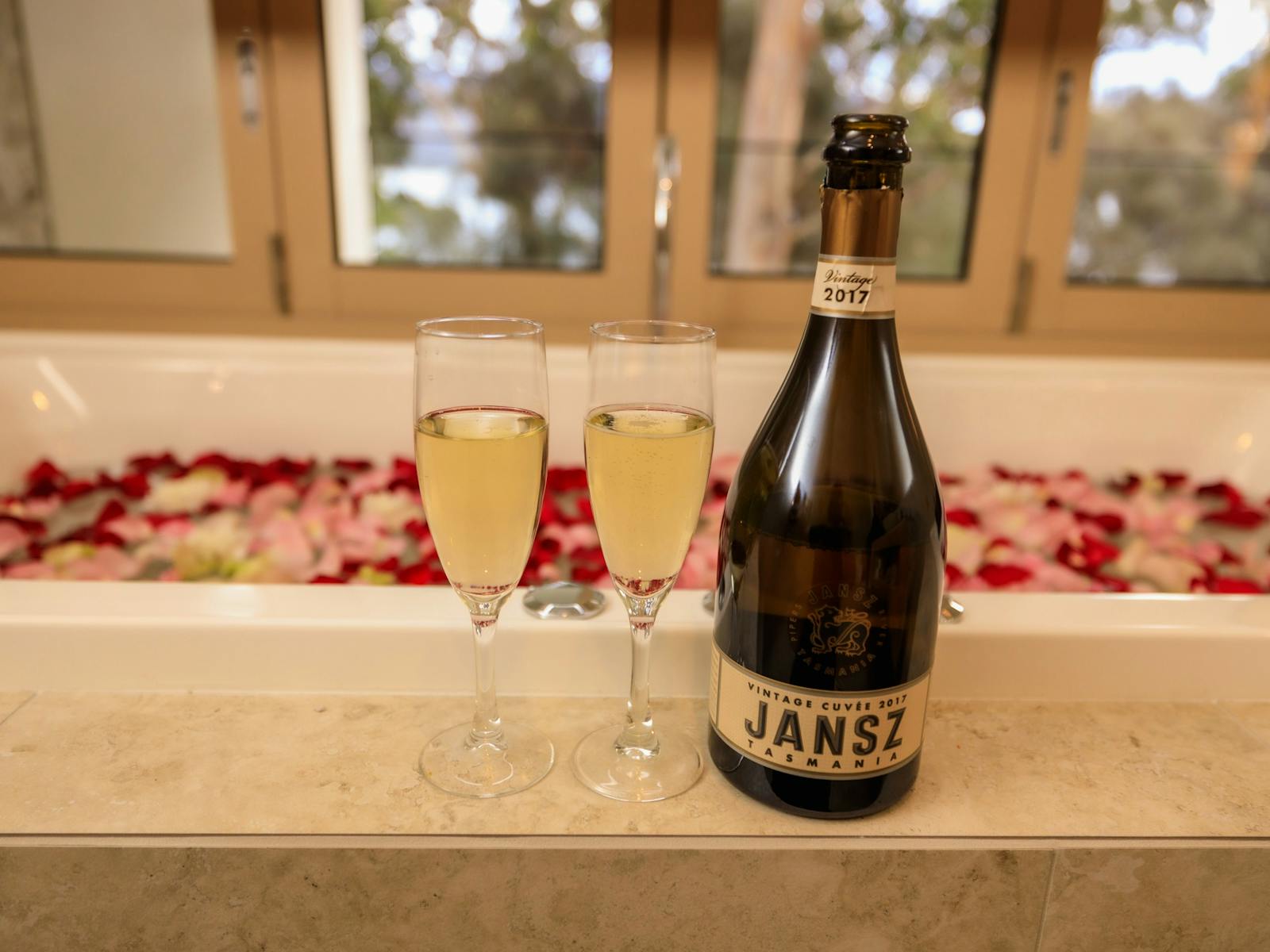 Bath filled with rose petals, champagne bottle and 2 glasses, ocean view through window.