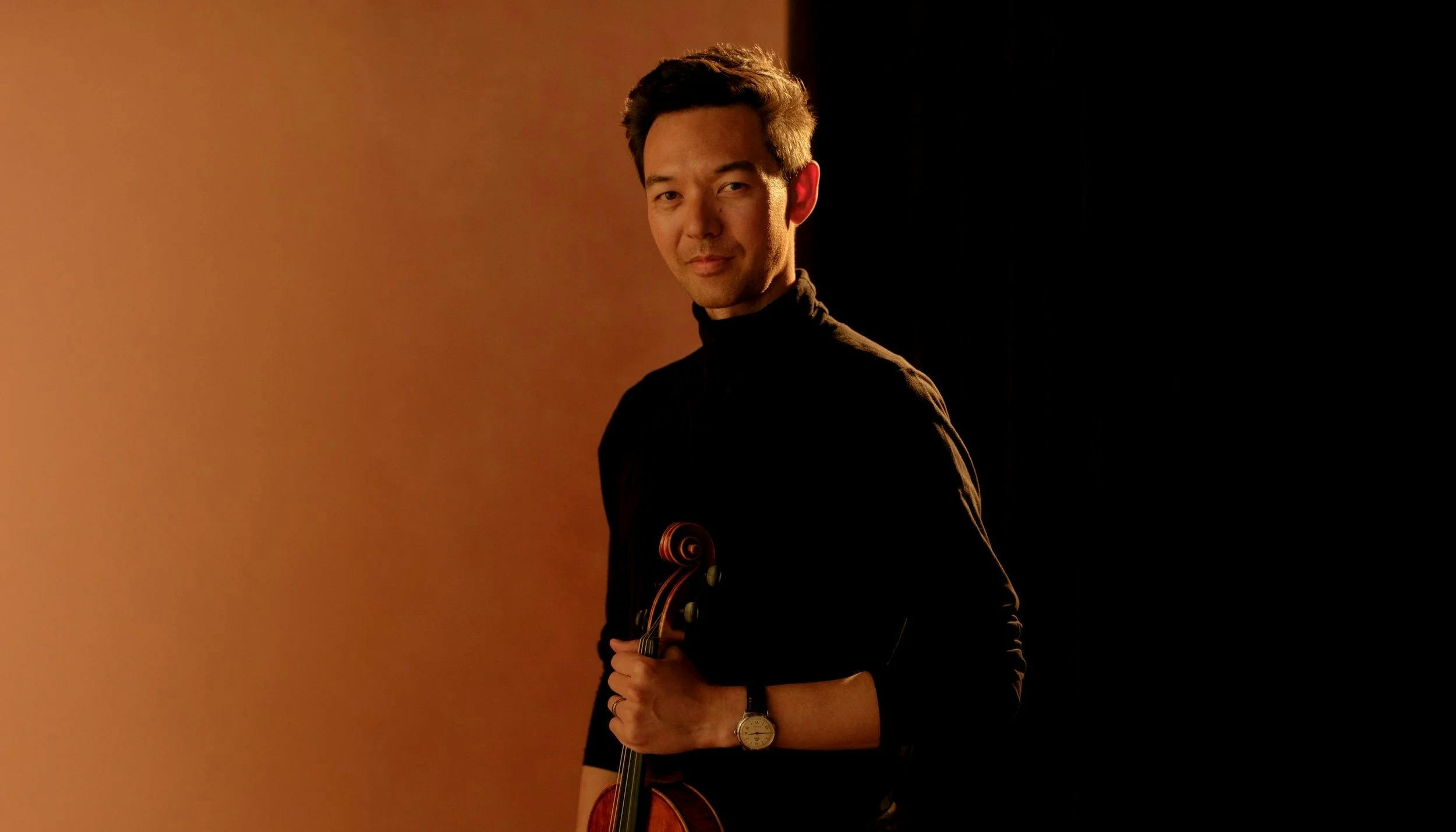 A person stands holding a cello against a warm, brown background.