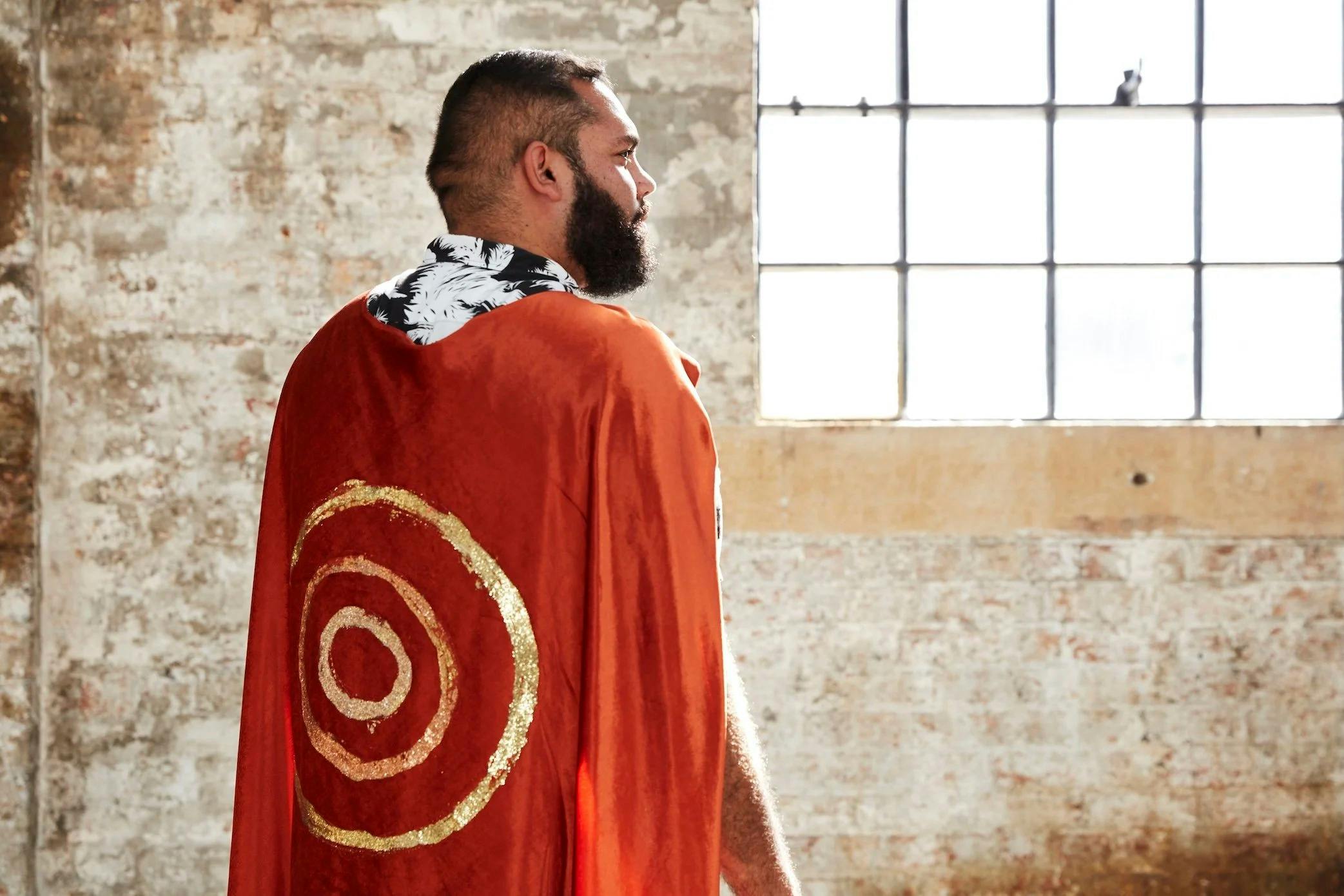 A man stand away from camera with a red cape and gold circles painted on it