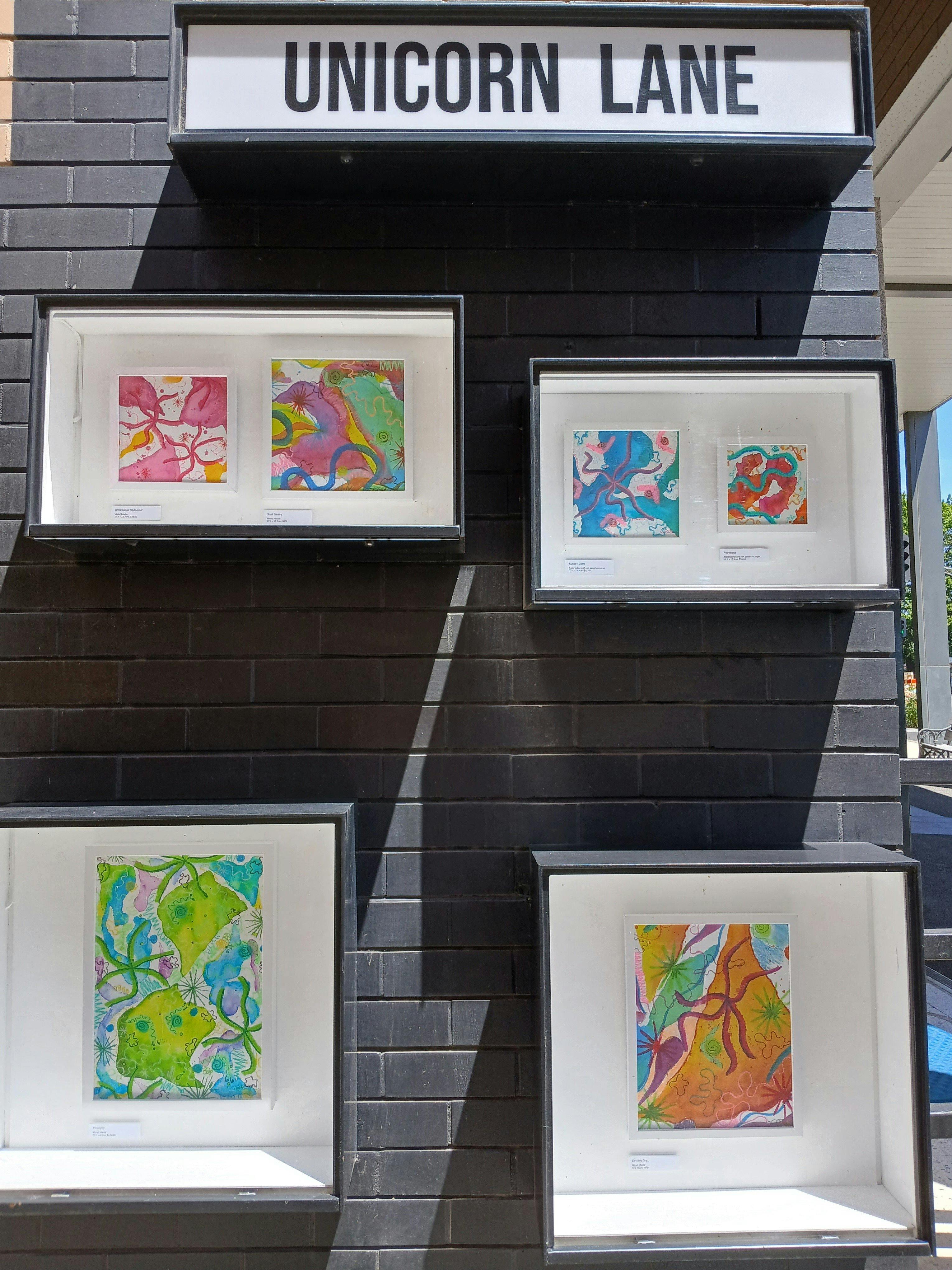 Two outdoor gallery boxes showing four pieces of brightly coloured abstract paintings.