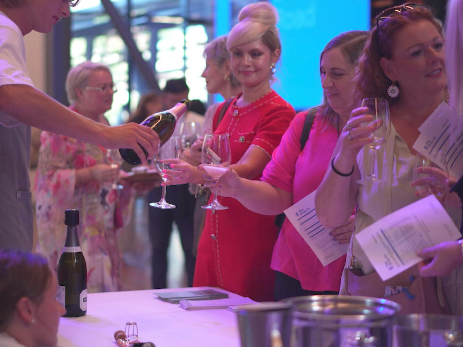 A server pours wine for women in bright pink outfits, holding glasses