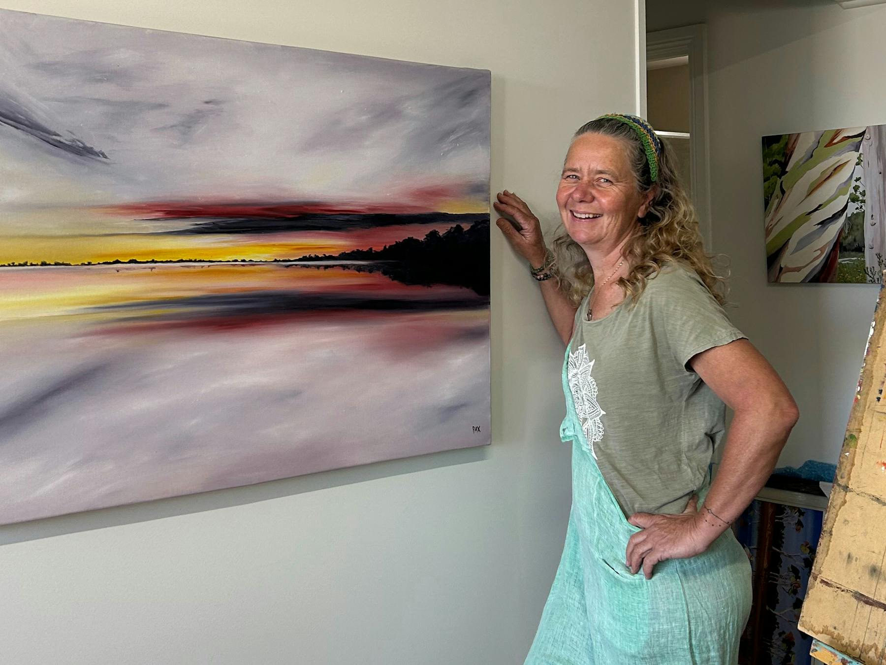 Artist Caroline Packer Pax with her painting Sunset Beach over Lake Lady standing with her painting