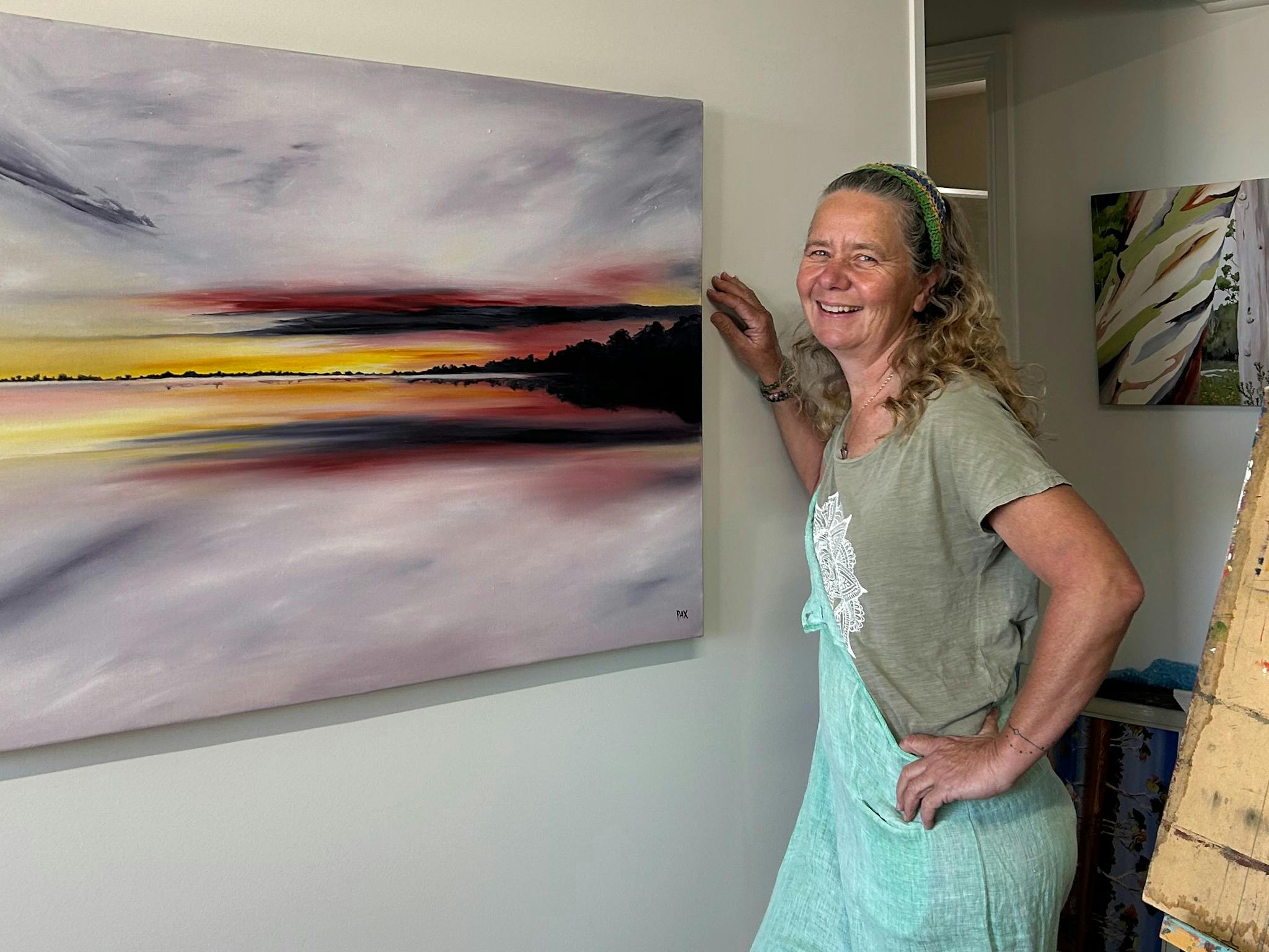 Artist Caroline Packer Pax with her painting Sunset Beach over Lake Lady standing with her painting