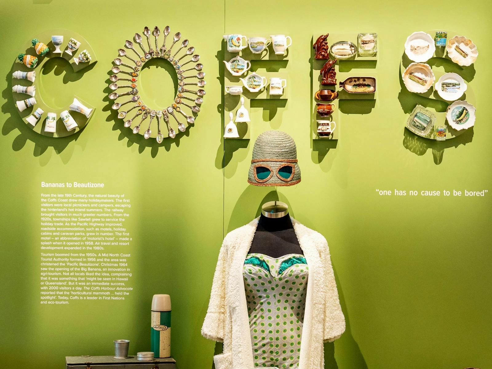 Lime green gallery wall spelling COFFS with crockery, above a mannequin in patterned dress.