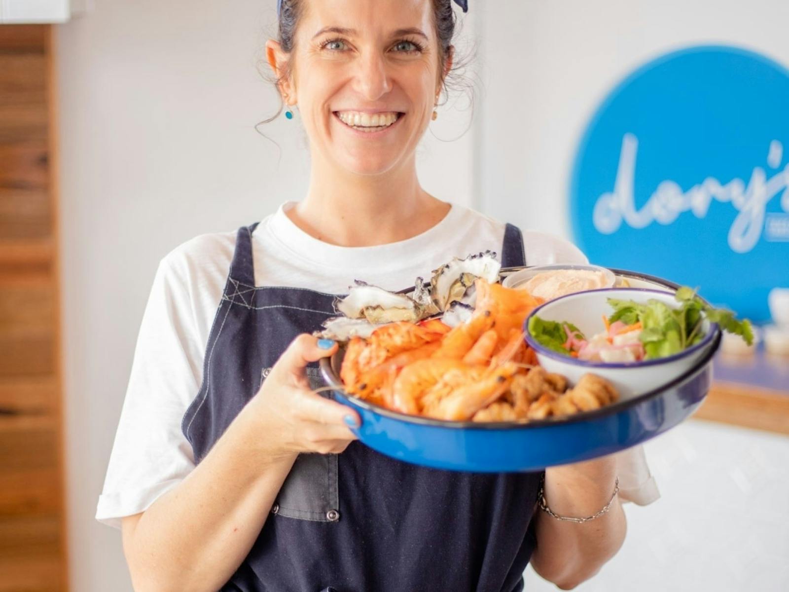 A lady smiling, wearing an apron and holding a large plate of seafood