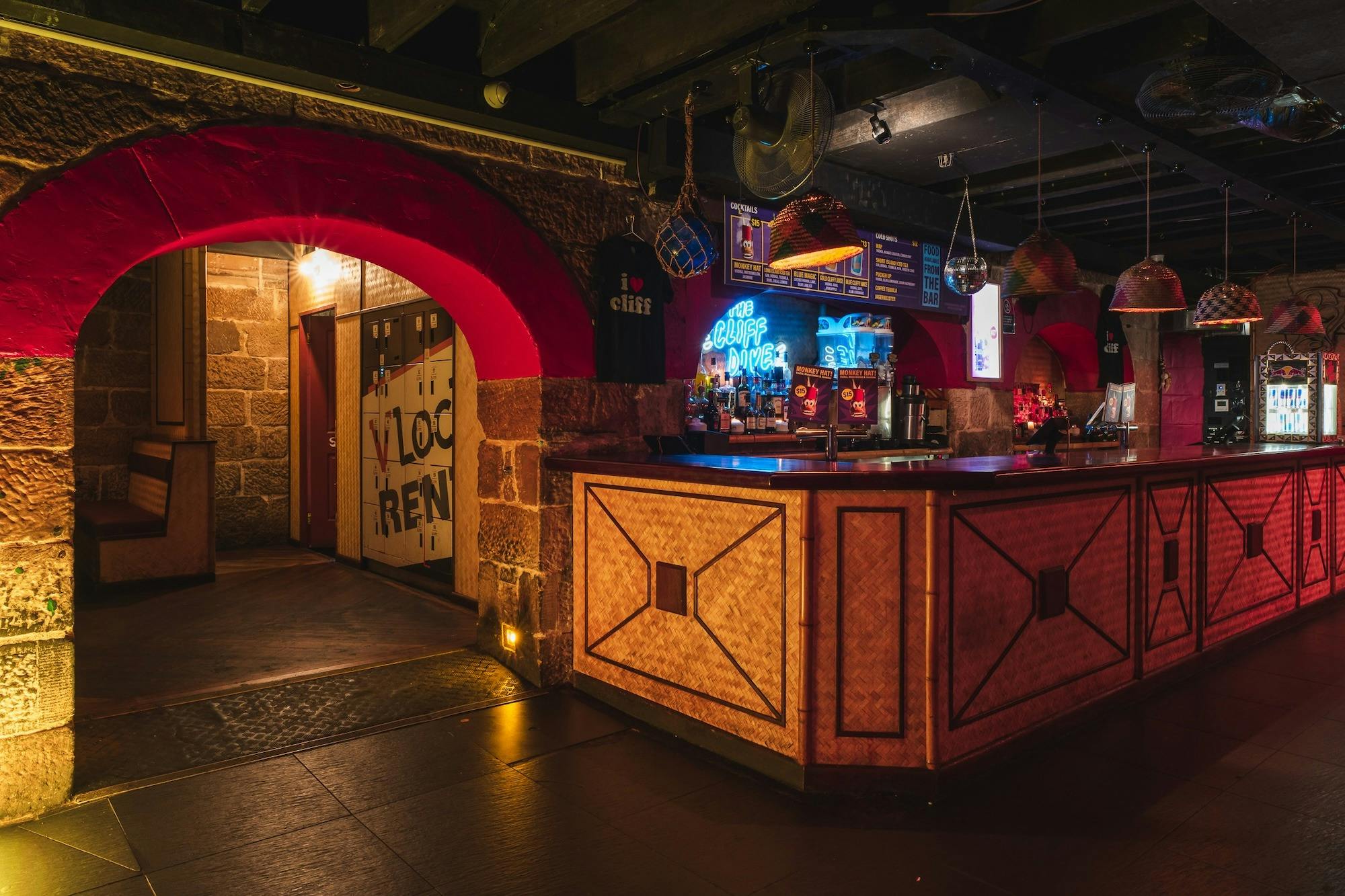 A nightclub bar area with woven wood paneling and warm lighting. A neon sign reading The Cliff Dive