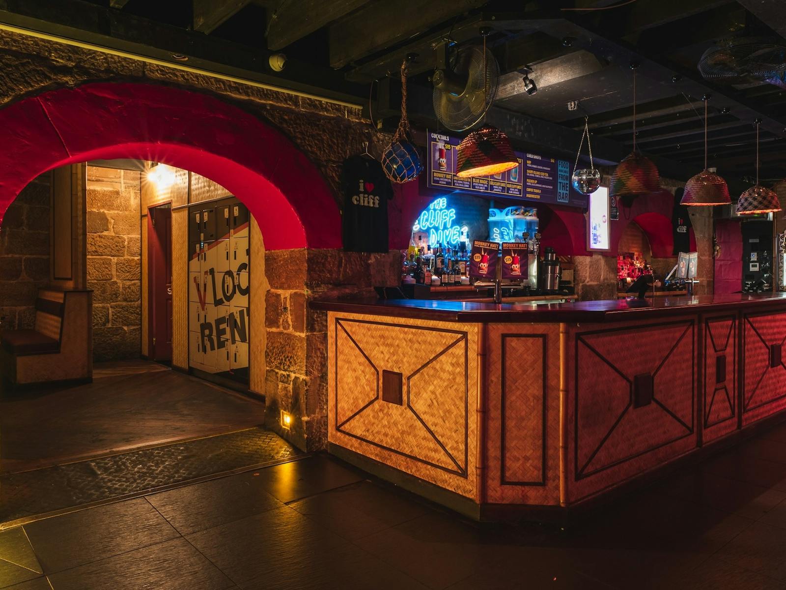 A nightclub bar area with woven wood paneling and warm lighting. A neon sign reading The Cliff Dive