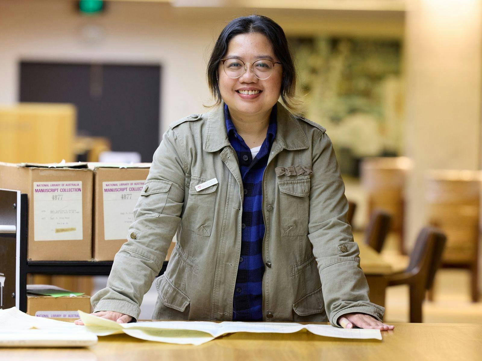 A woman in a green jacket stands leaning on a table covered with papers