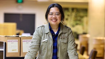 A woman in a green jacket stands leaning on a table covered with papers