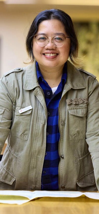 A woman in a green jacket stands leaning on a table covered with papers