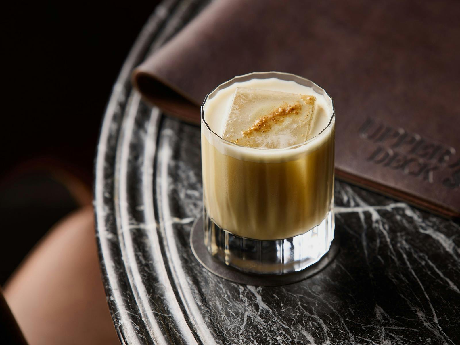 A creamy looking cocktail sat on a dark table, with a leather menu to the side of it.