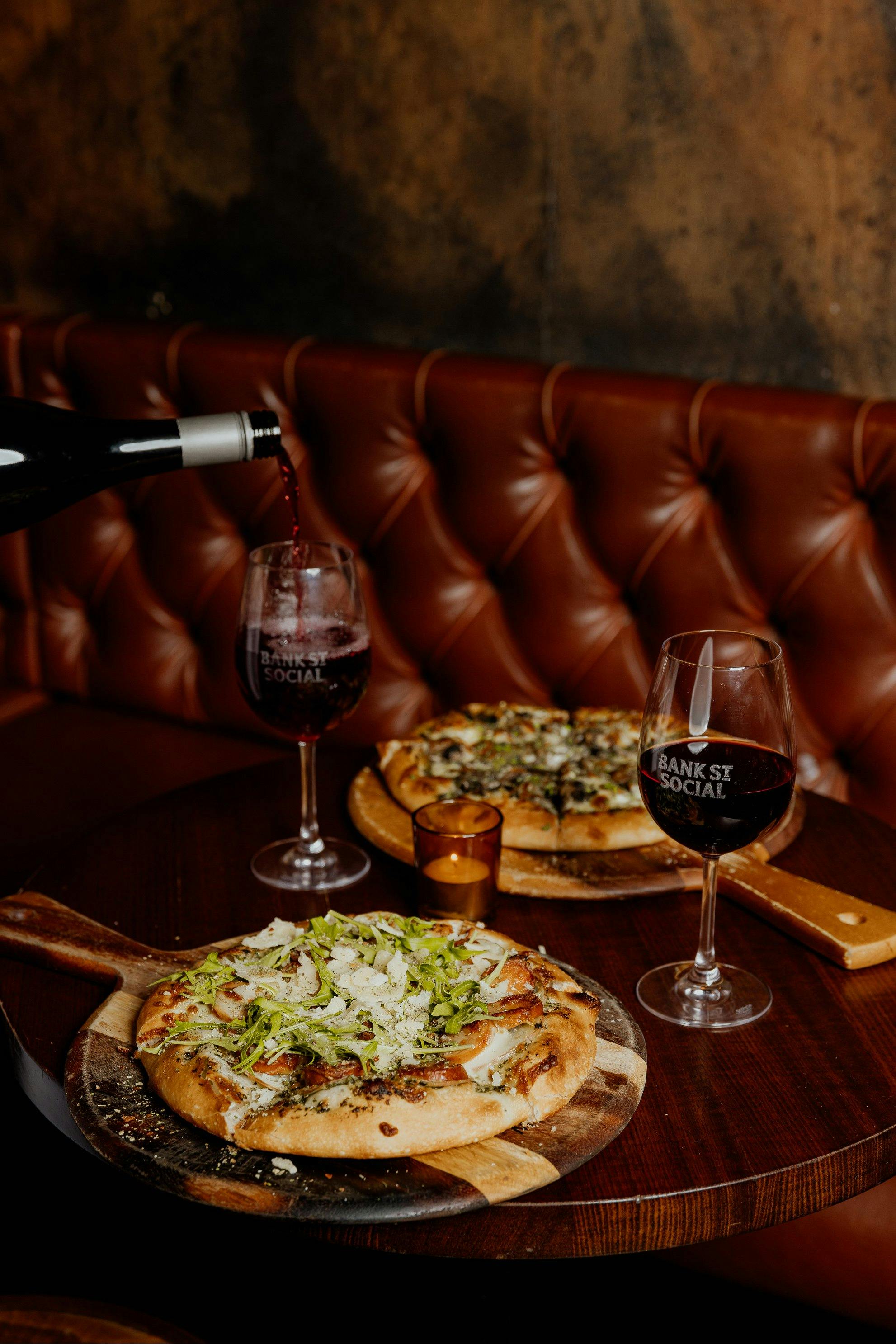 Two Pizza's placed on a booth table with red wine being poured into two wine glasses.