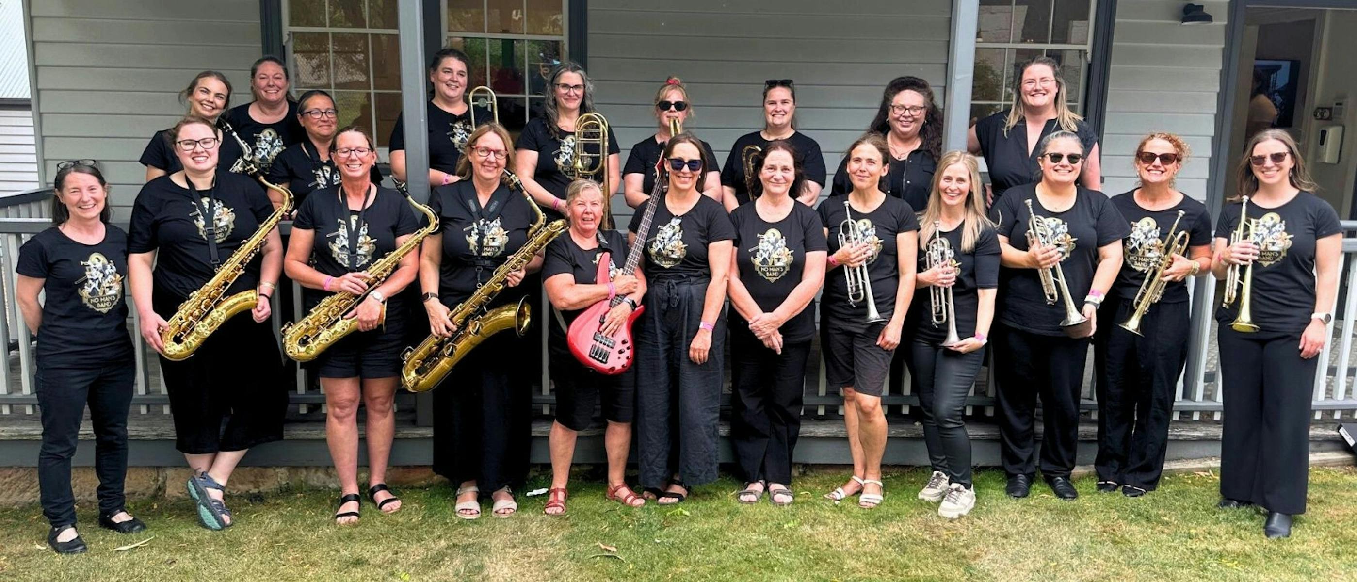A group of women holding trumpets, saxaphones and guitar stand outside together, smiling