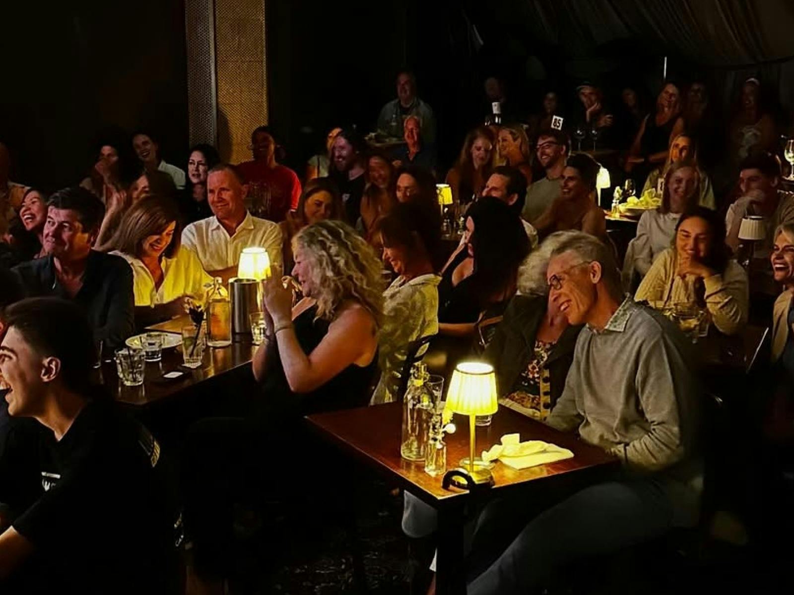 A large audience seated at tables with small lamps, smiling and laughing at a comedy show