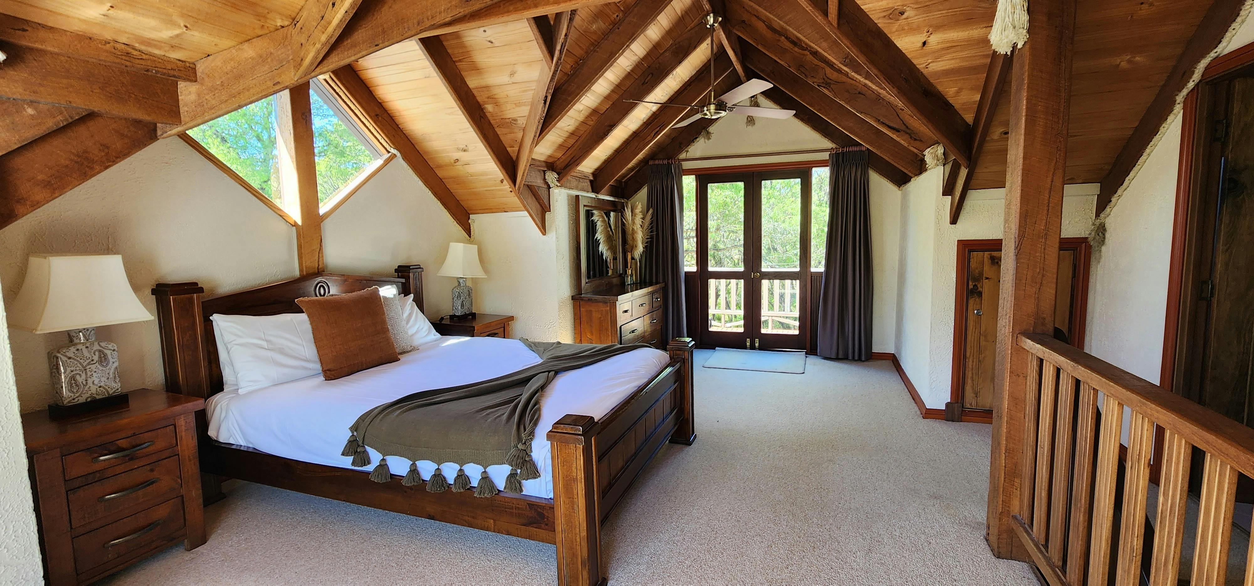 Large cosy cabin bedroom, surrounded by trees, with pitched wood ceilings and large window above bed