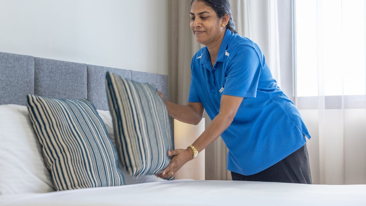 A woman with dark hair wearing a blue polo shirt placing a cushion on a bed.