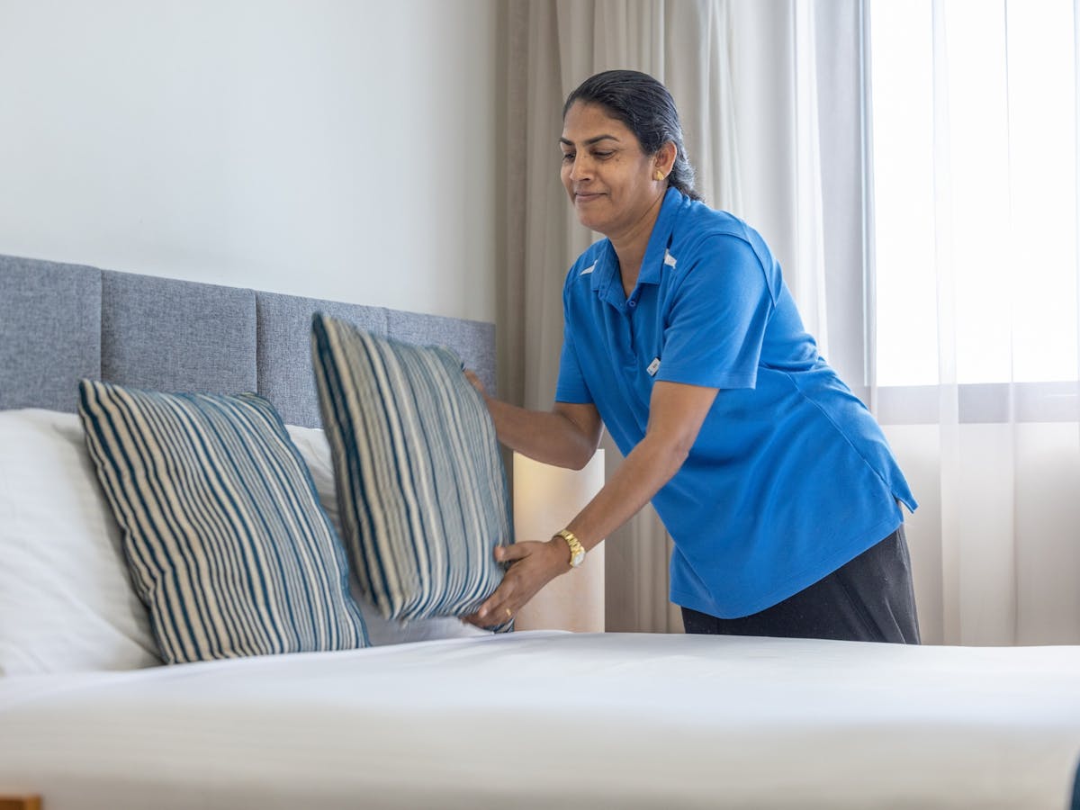 A woman with dark hair wearing a blue polo shirt placing a cushion on a bed.