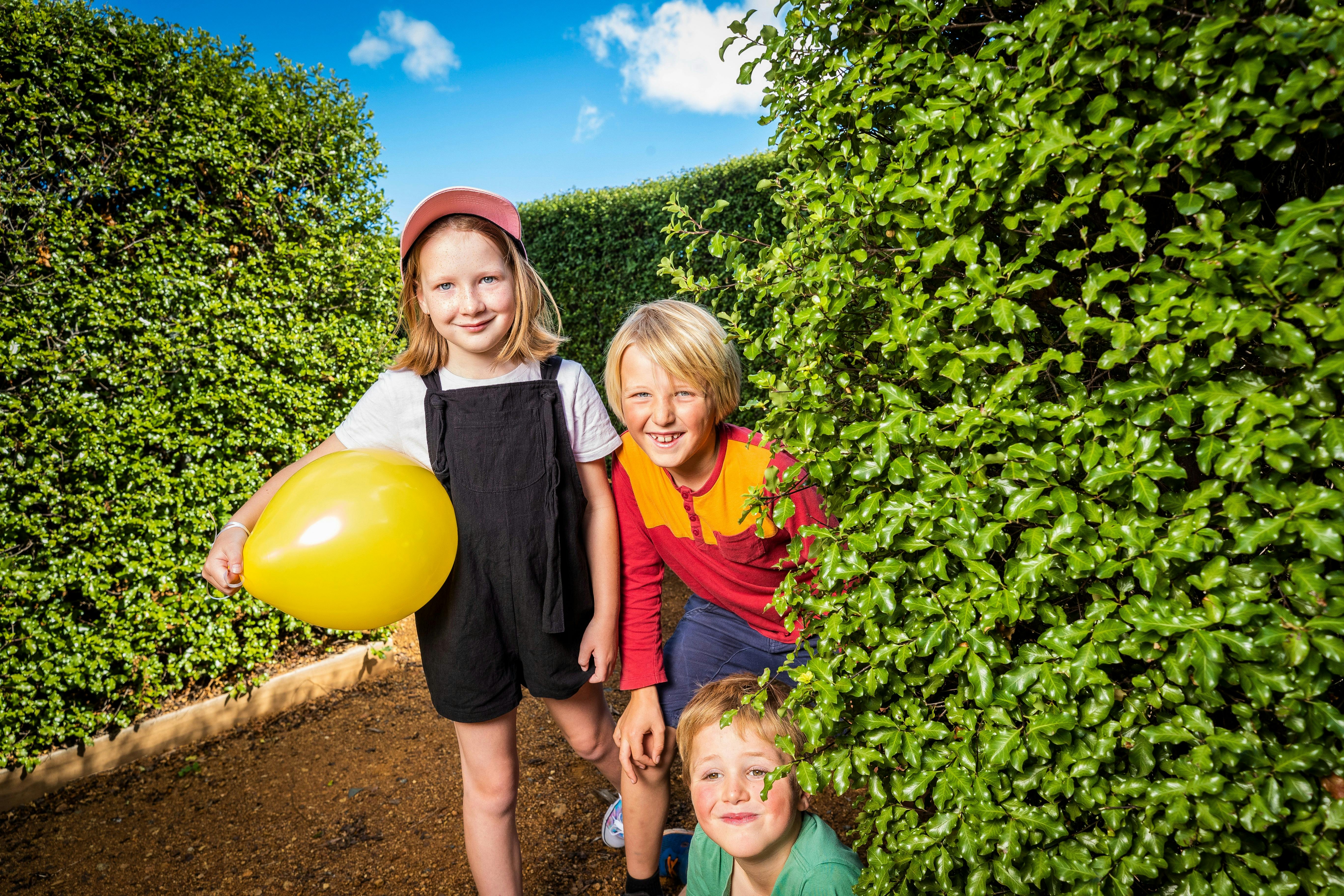 Three children in a lush green hedge maze at Amaze Richmond. Girl is holding a yellow balloon.
