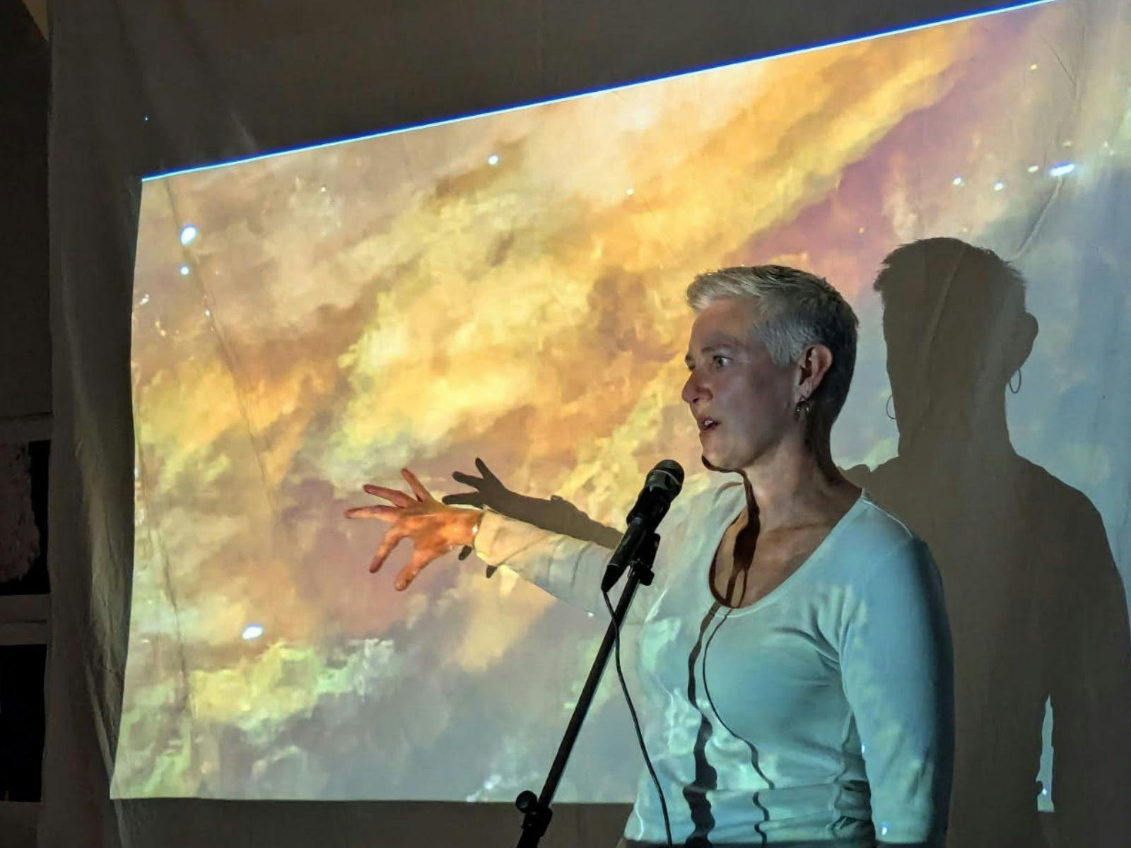 A woman stands at a micorphone in a projection of yellow and rust-coloured moving water
