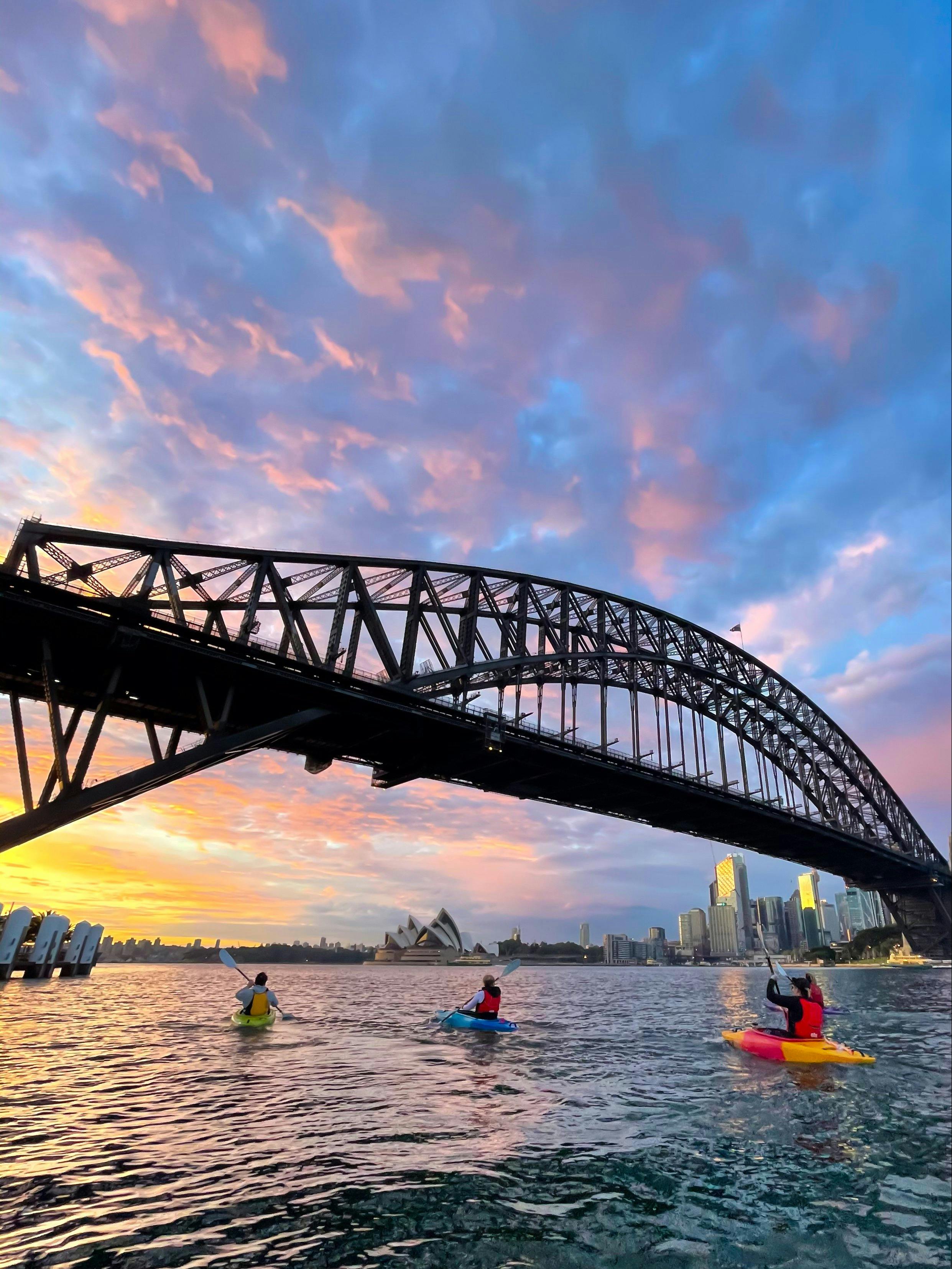 Three kayakers looking up at Sydney Harbour Bridge and across at Sydney Opera House.