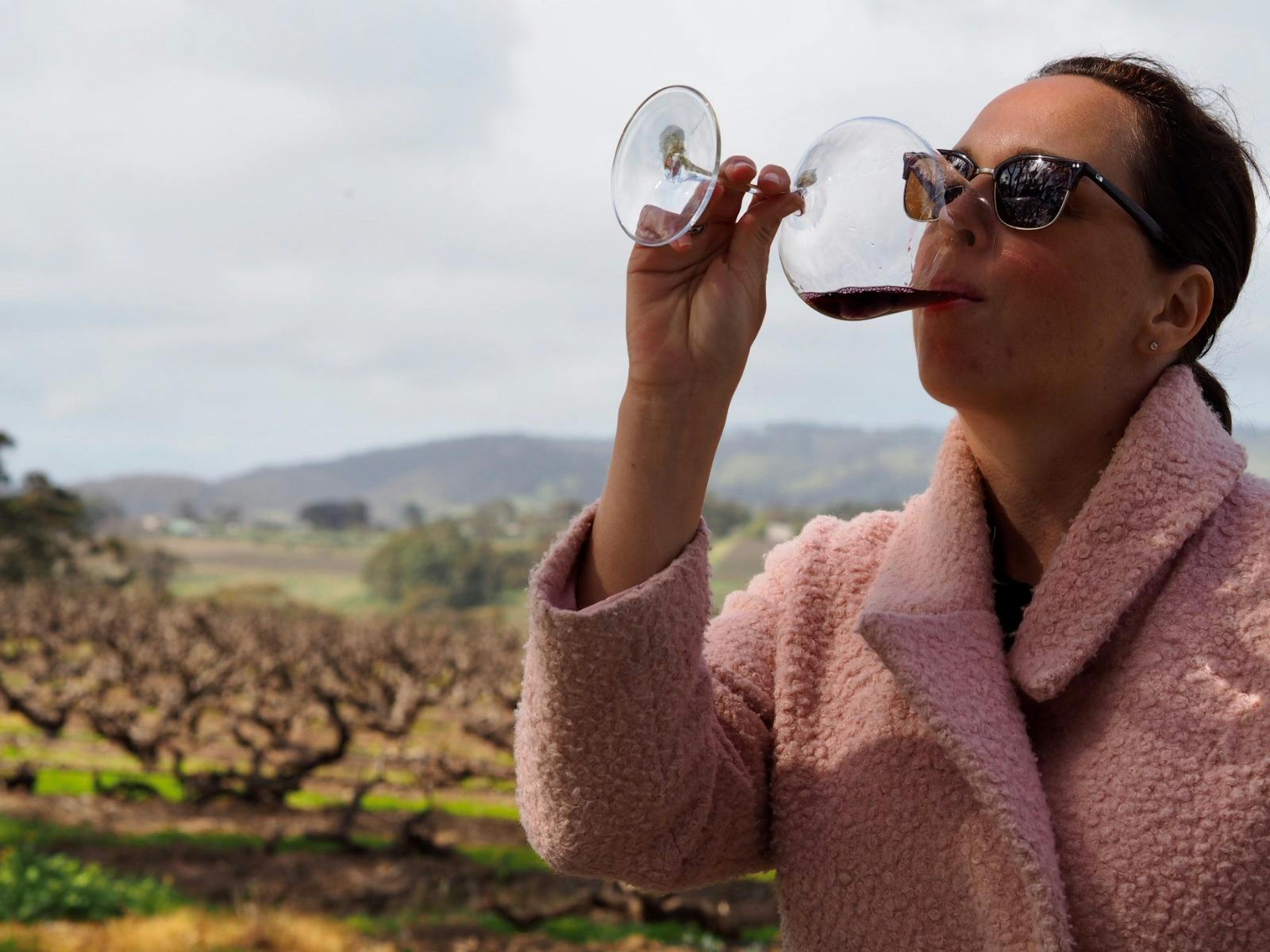 A woman takes a sip of Aphelion Wine with a background or bare wintery grape vines and green hills