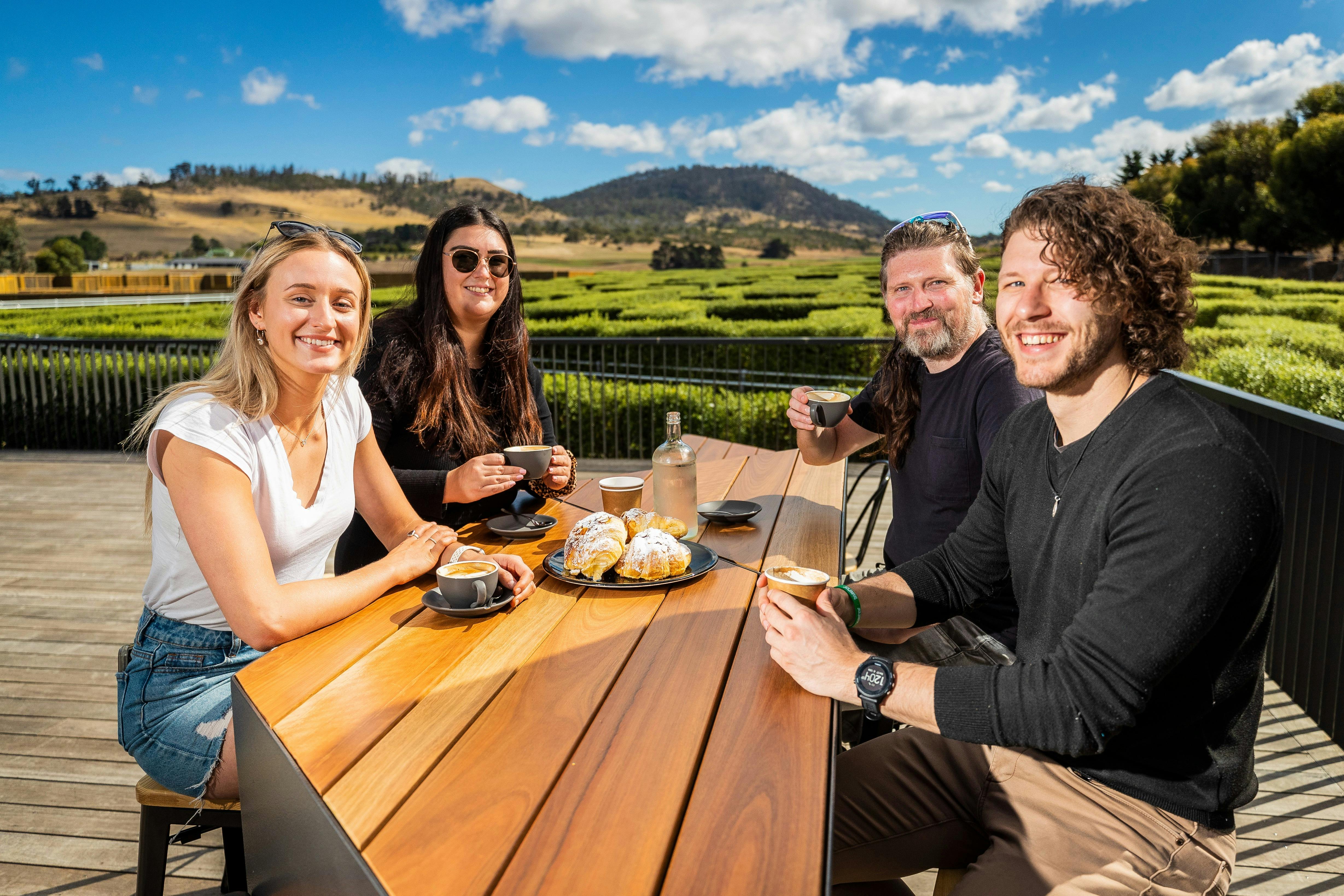 Two women and two men drinking coffee at a table. View of hedge maze in the background.