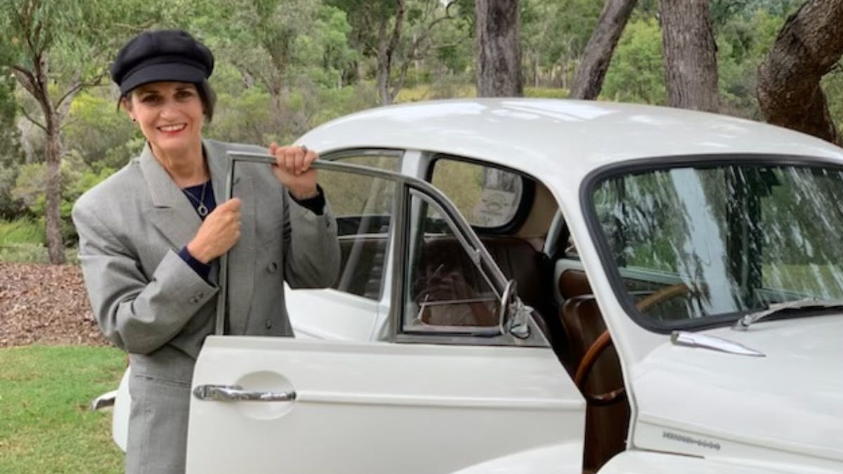 Woman wearing hat, stands next to vintage car with the door open, she is smiling