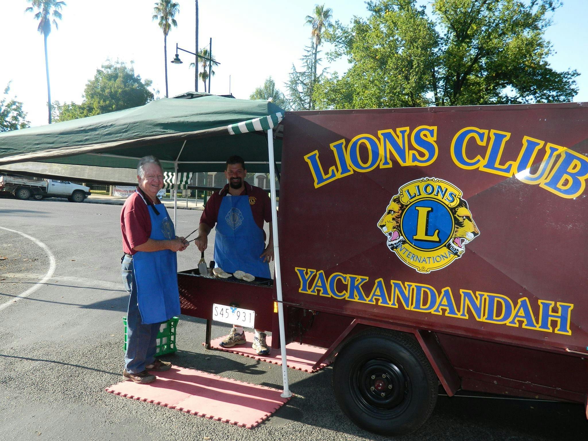 BBQ trailer with 2 men wearing blue aprons, cooking sausages and hamburgers