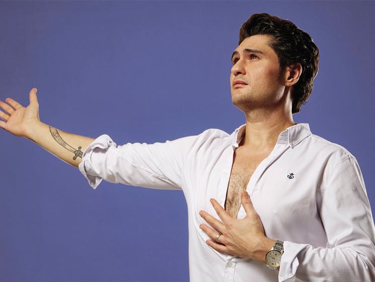 Young male adult wearing white shirt with his left hand on chest, right arm open. Background is blue