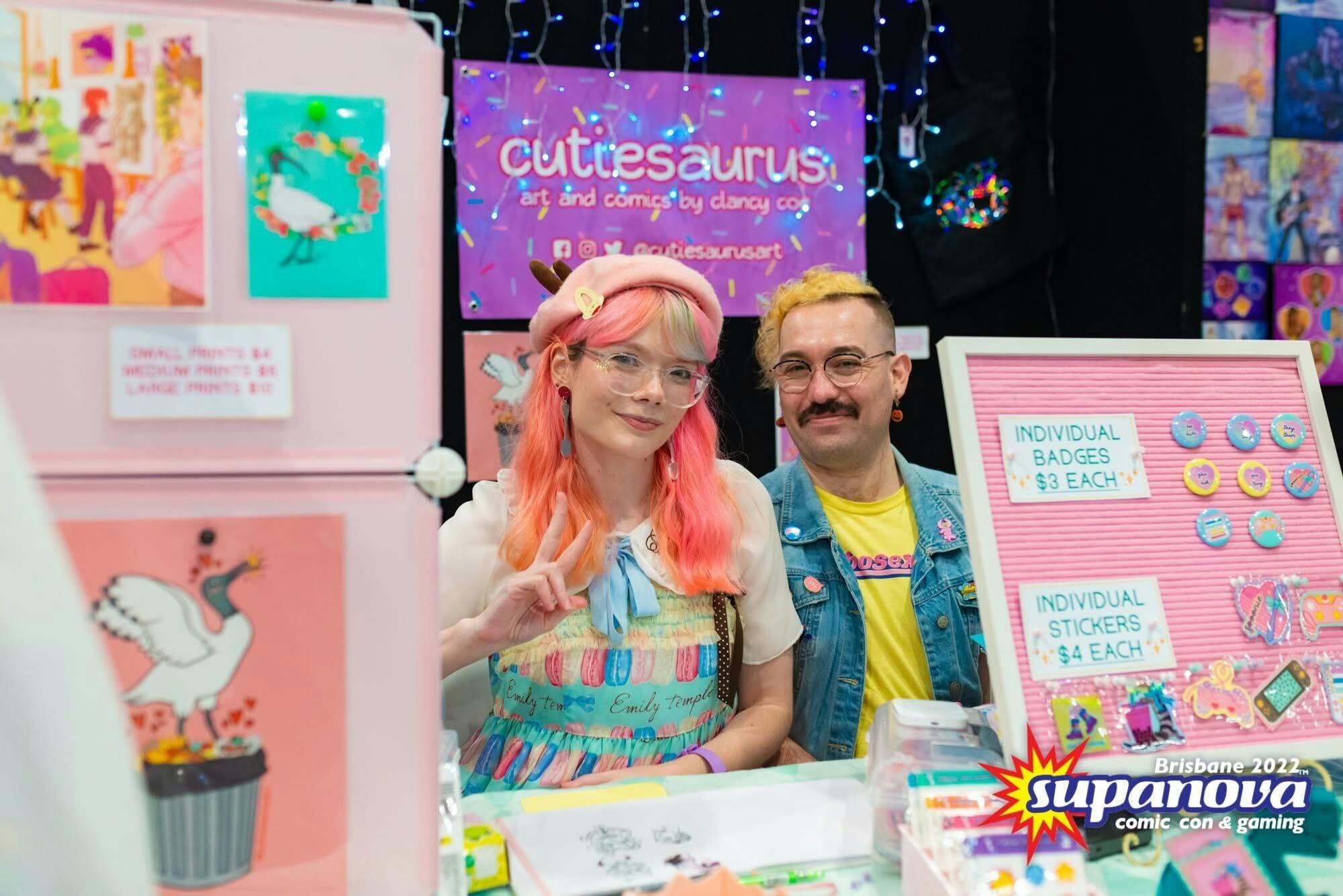 Two artists smile at their colourful booth for Cutiesaurus at Supanova Brisbane