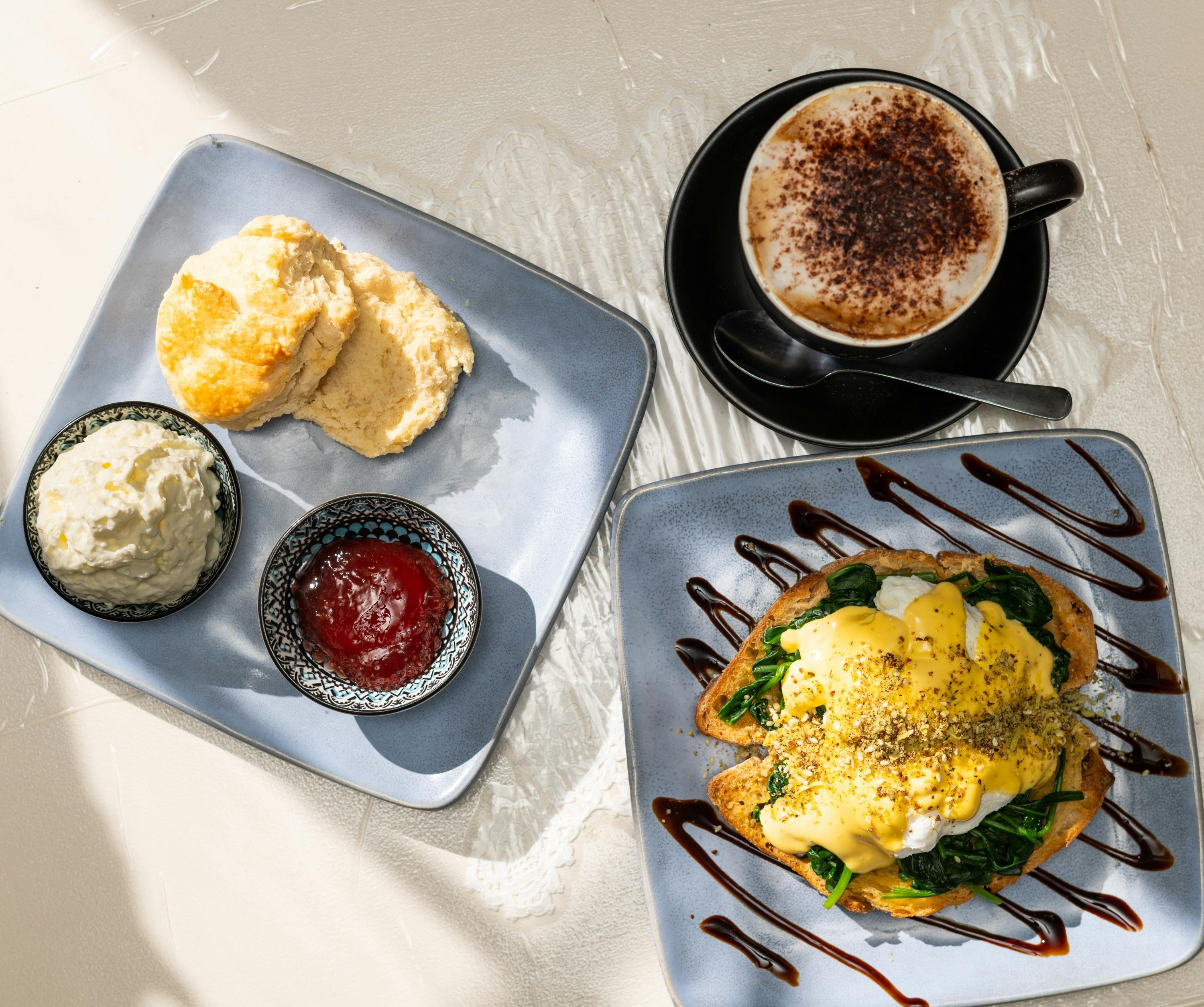 Plate of scones, cream and jam. Beside it, a plate of eggs benedict and a cappuccino.