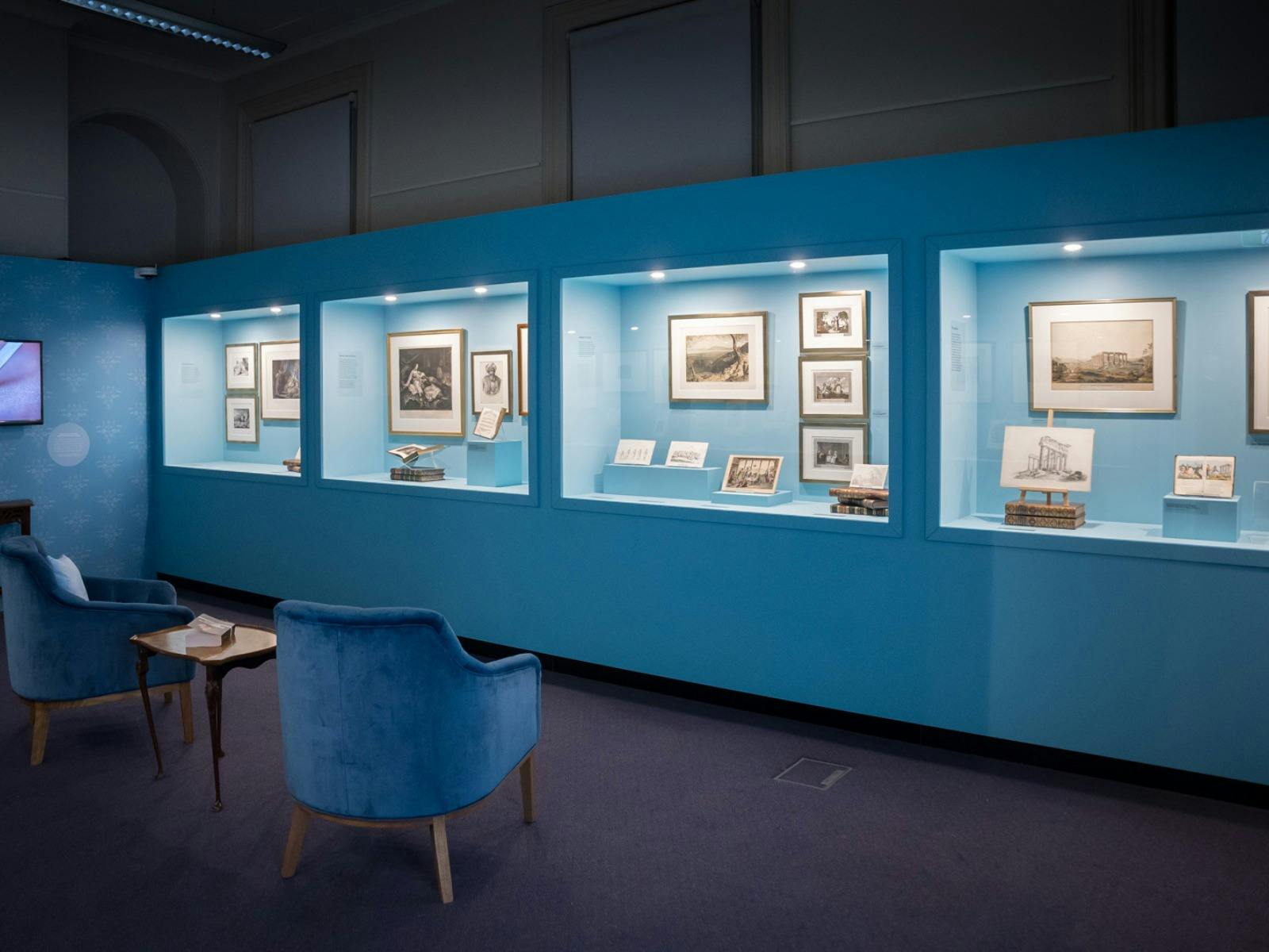 Four cabinets containing an array of artworks in a blue gallery with an armchair in foreground