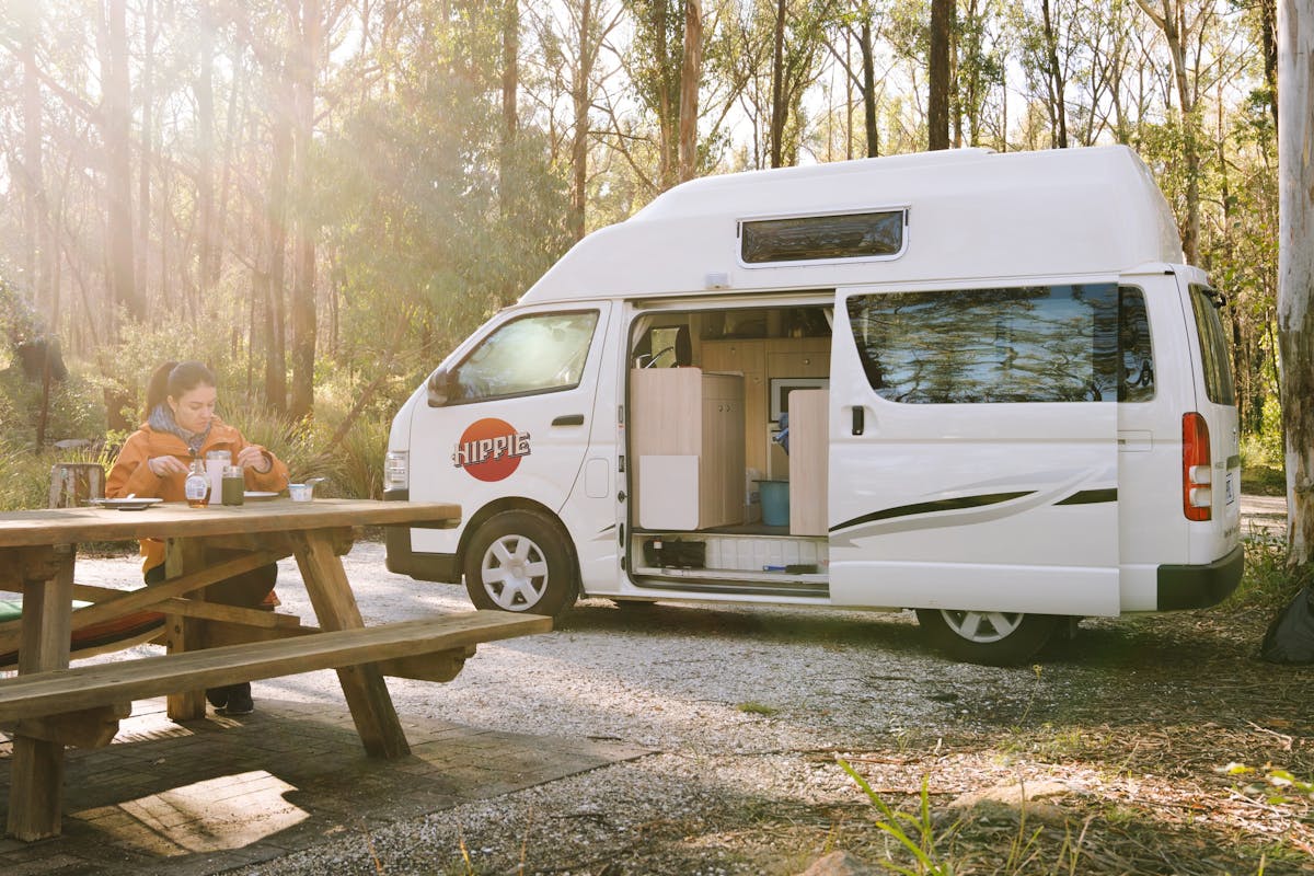 A campervan is parked next to a picnic table where a woman is eating breakfast