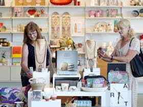 Two women browse items in a museum shop; in the foreground is a shop display table with ceramics